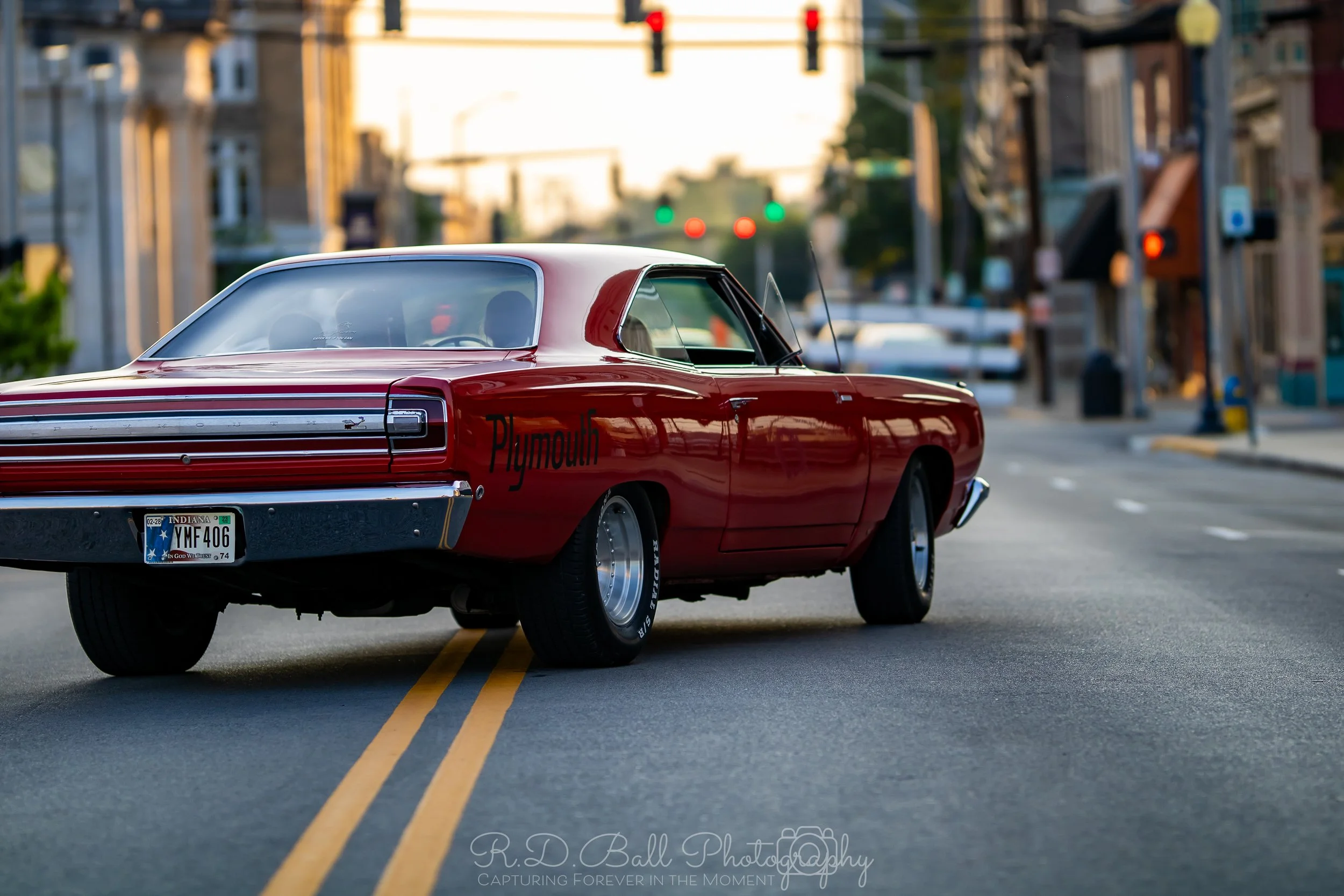 A vintage red Plymouth car parked on an urban street with buildings and traffic lights in the background.