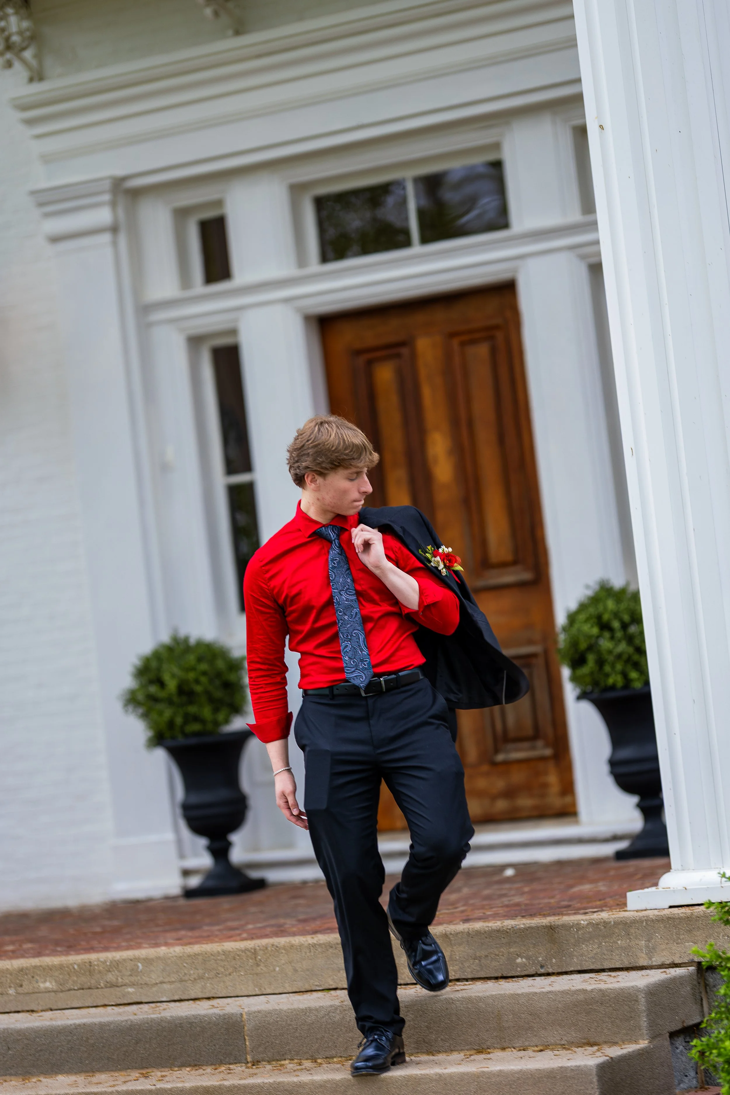 A young man dressed in a red shirt, black pants, and a tie, holding a jacket over his shoulder with a boutonnière, walking down the steps in front of a large house with a wooden door and potted plants.