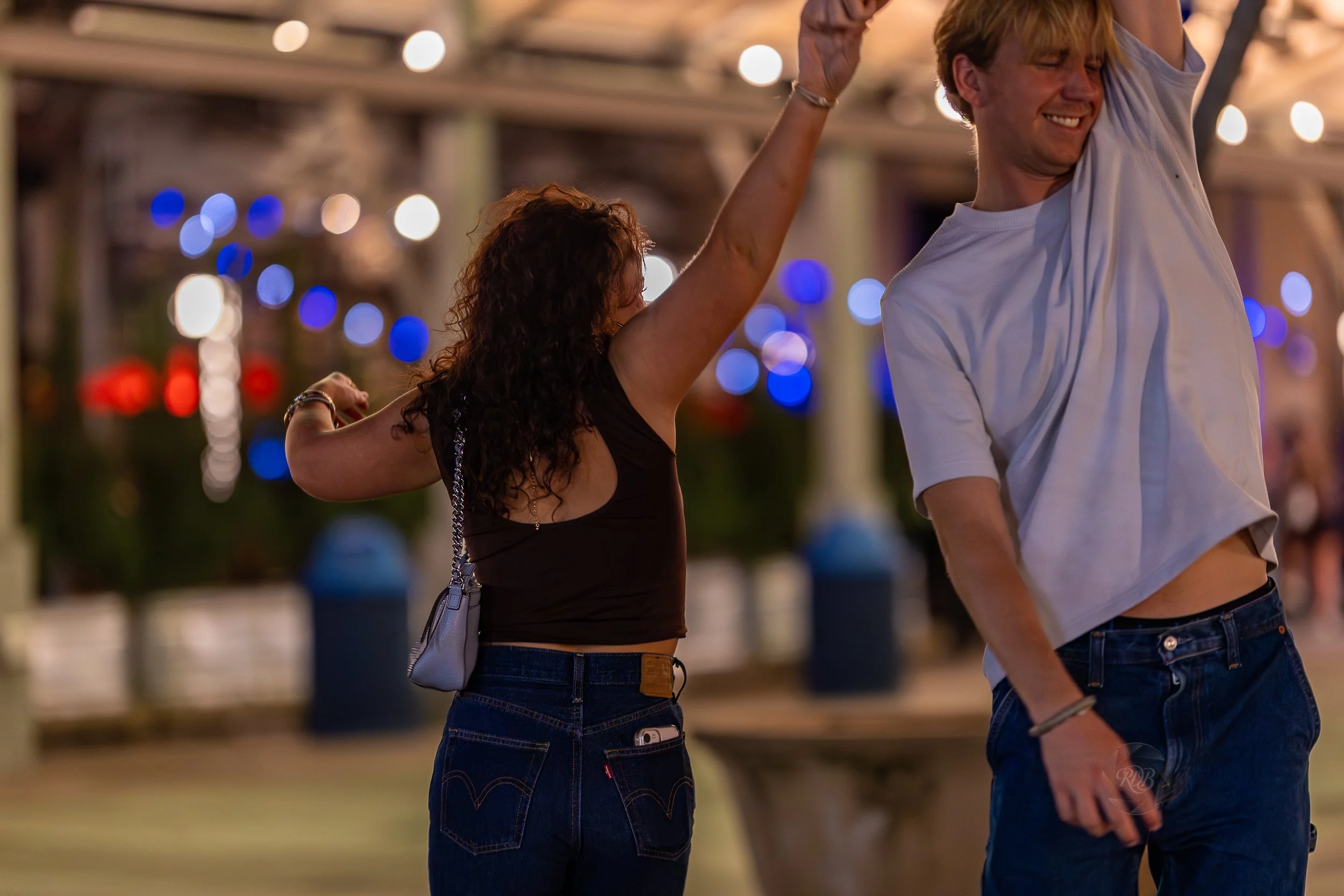 A man and woman dancing at night in a lively, festive outdoor environment with colorful string lights and blurred background.