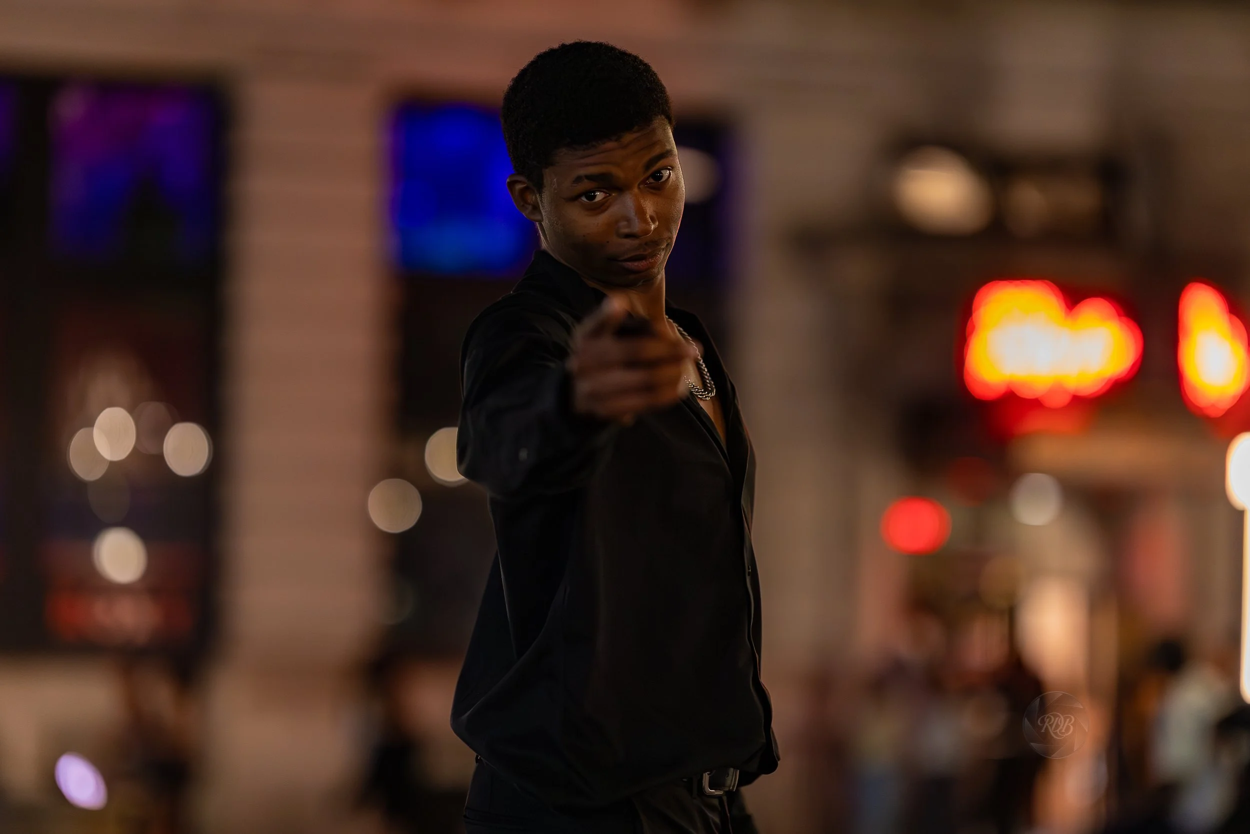 A young man in a black shirt points towards the camera in a dimly lit city street at night, with blurred colorful neon signs in the background.