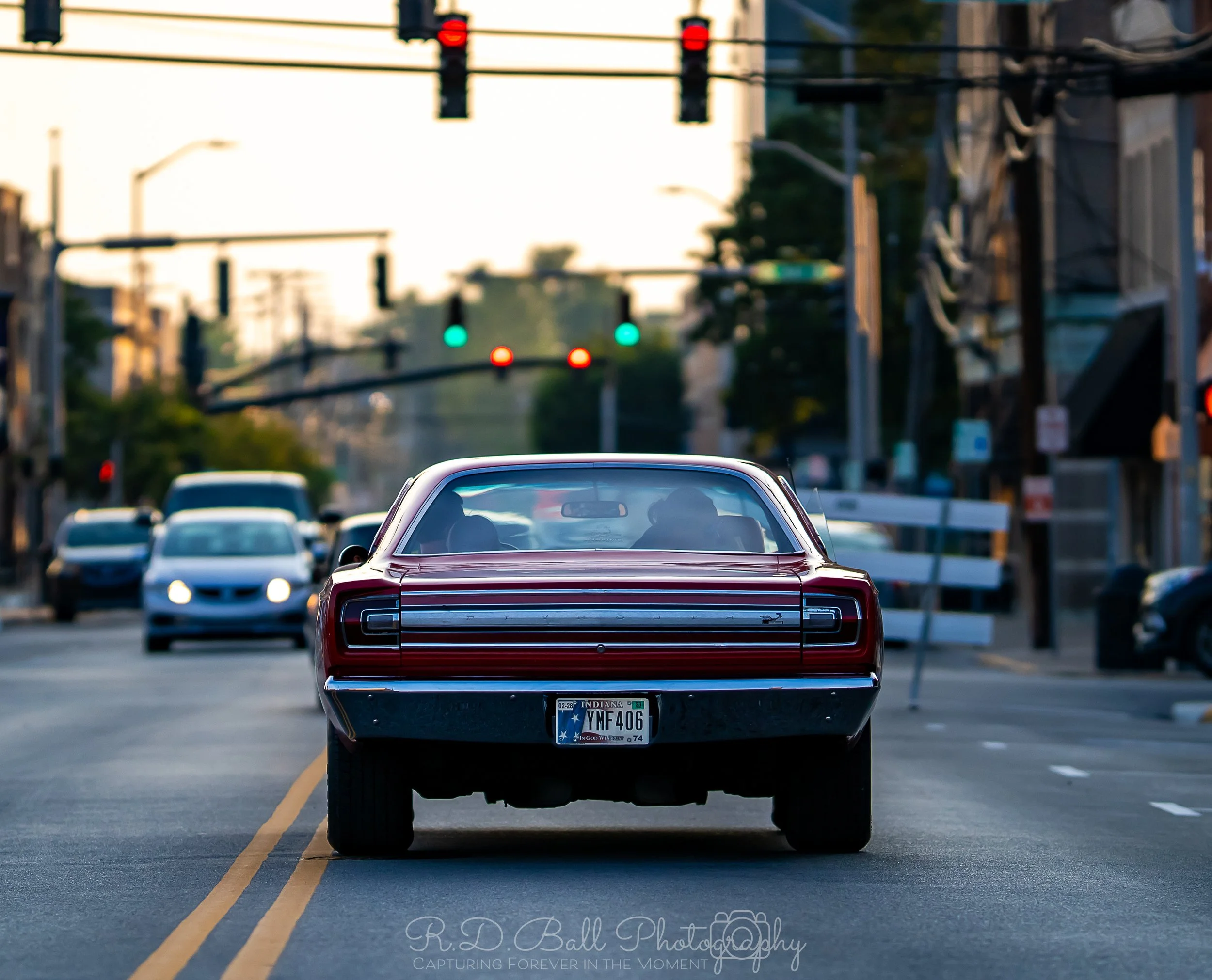 A classic red muscle car driving down a city street at dusk, with traffic lights overhead and other vehicles ahead.