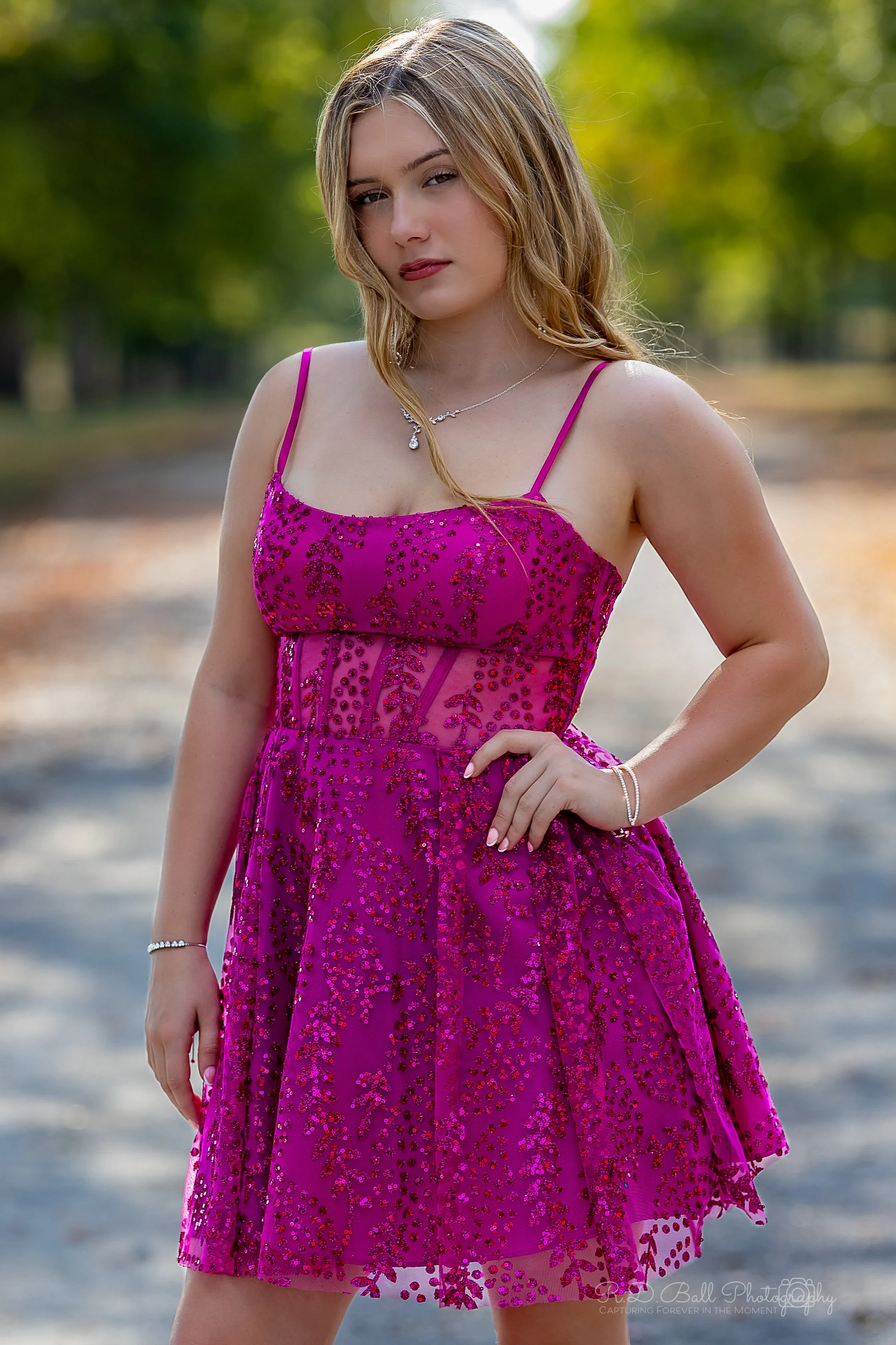 A young woman in a pink, sequined dress standing outdoors on a dirt path with trees in the background.