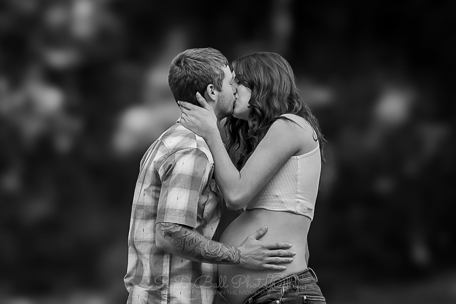 A black and white photo of a couple kissing. The man has tattoos on his arm and is wearing a plaid shirt. The woman has long wavy hair and is wearing a sleeveless crop top. The woman’s hands are on the man’s face, and the man’s hand is on her waist.