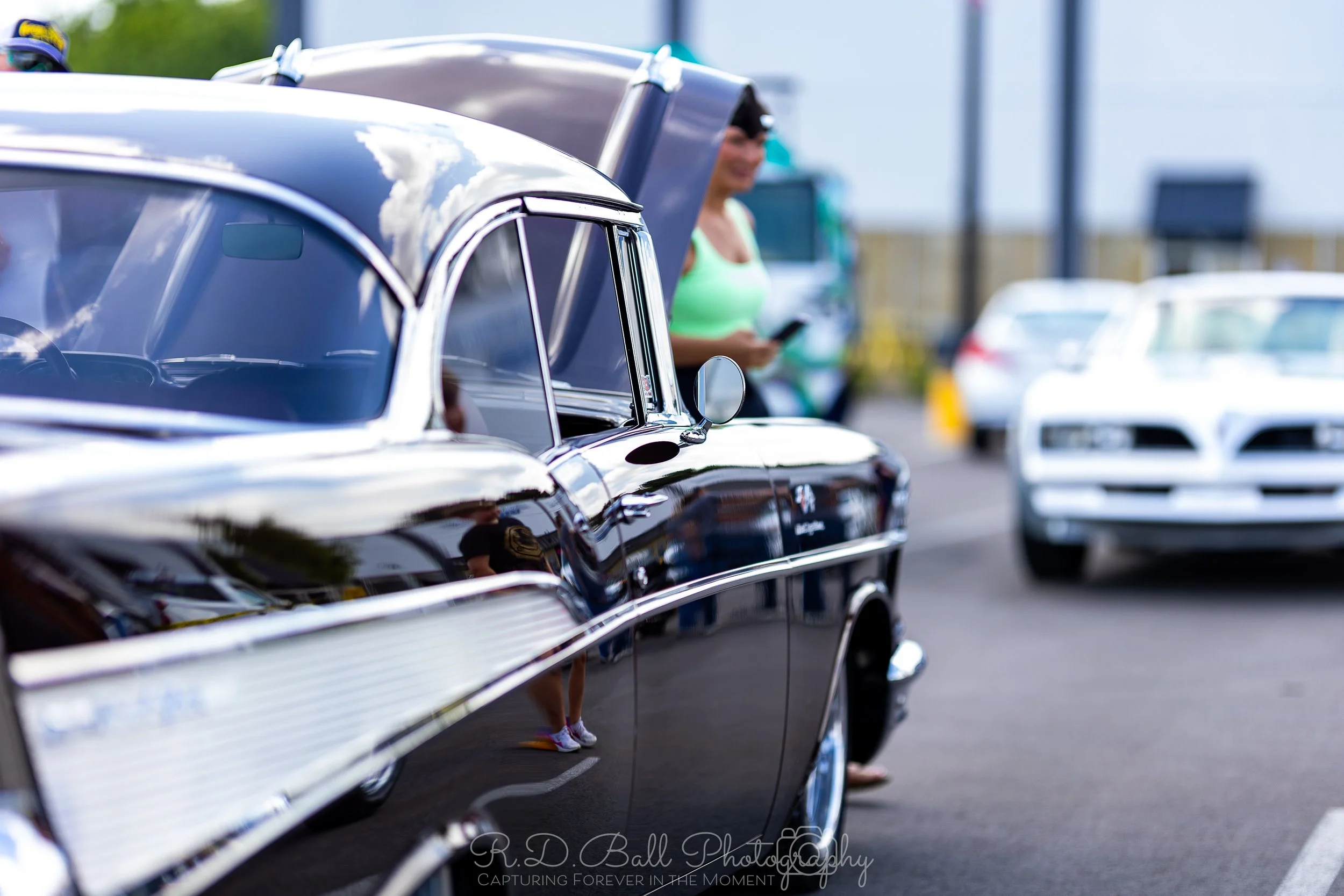 Close-up of a black classic muscle car in a parking lot, with a woman in a neon green top and sunglasses standing behind it, holding a phone. Other cars are visible in the background.