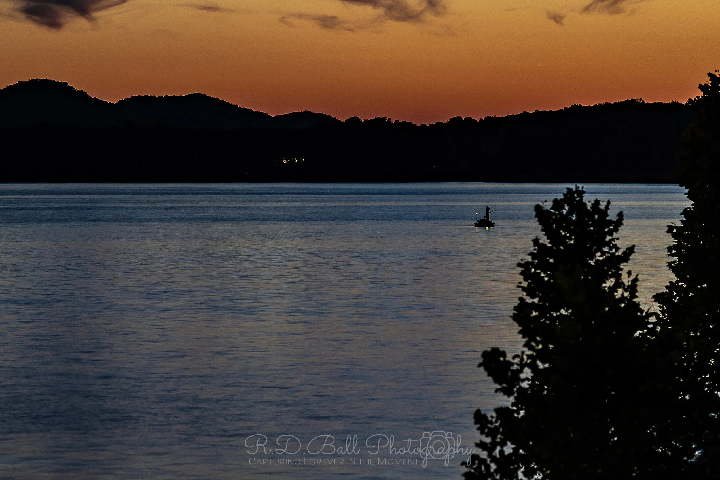 A peaceful lake at sunset with a small boat and person in the water, silhouetted against the colorful sky and mountains in the background, with dark trees in the foreground.