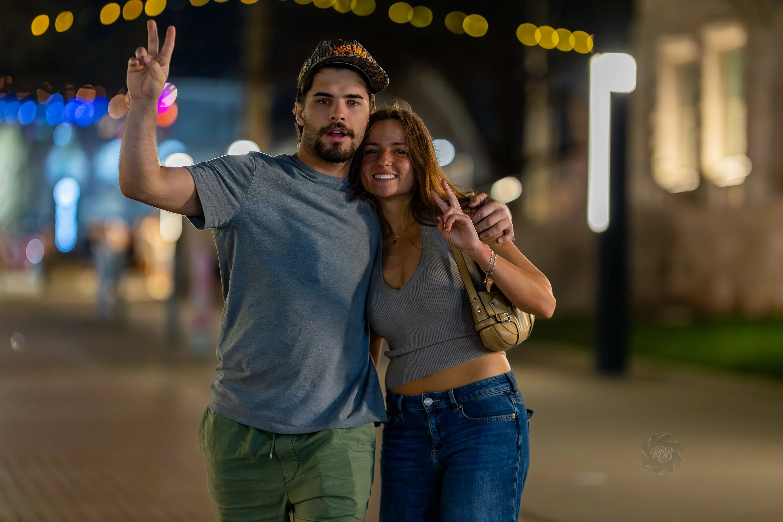 A young man and woman standing together at night on a city street, smiling and making peace signs with their hands, with blurred colorful lights in the background.