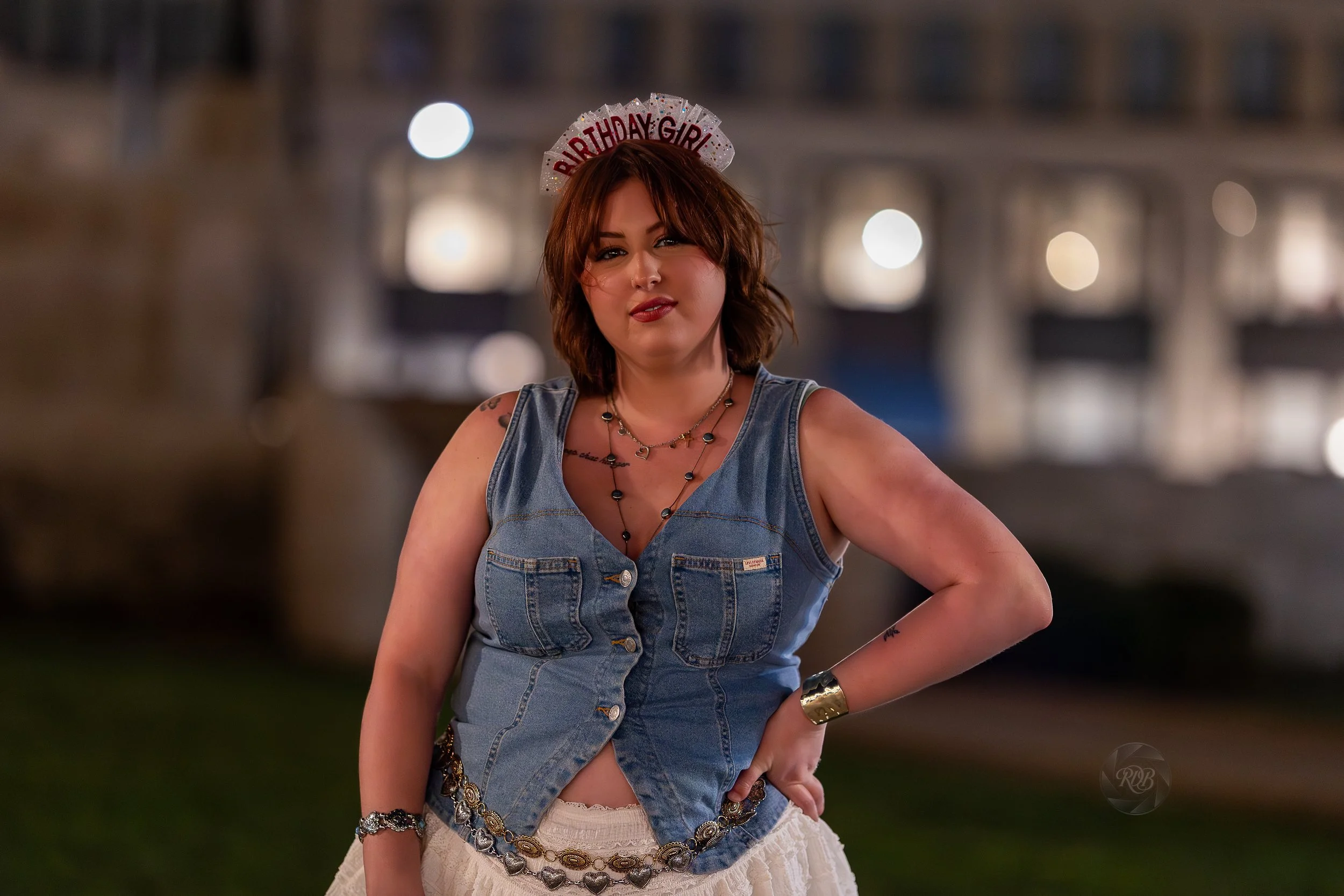 A woman with shoulder-length brown hair wearing a 'Birthday Girl' headband, denim vest, layered necklaces, bracelets, and a beige skirt, standing outdoors at night with a blurred cityscape background.