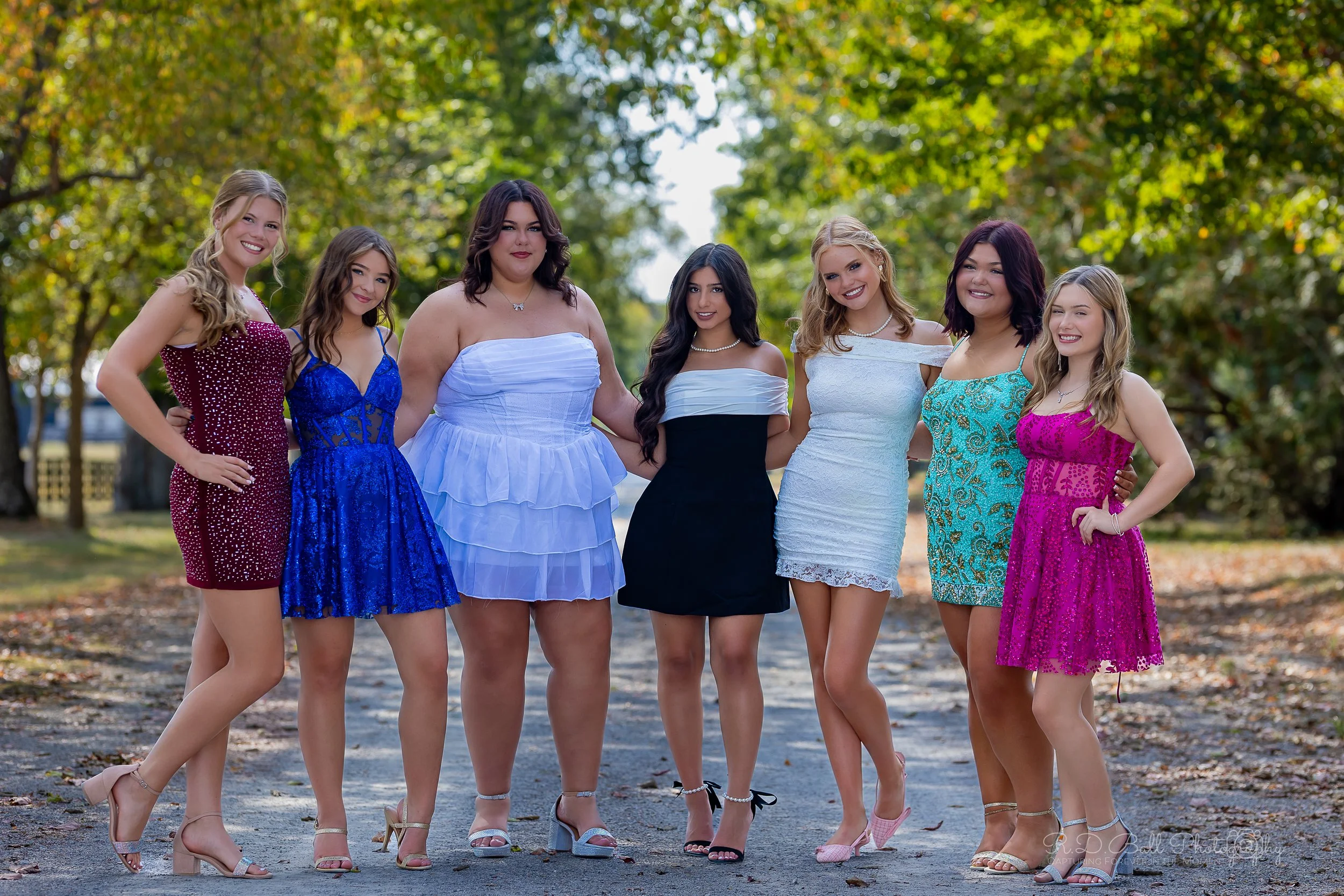 Group of seven young women standing outdoors on a dirt path surrounded by trees with fall foliage. They are dressed in colorful dresses with heels, posing for a photo and smiling.