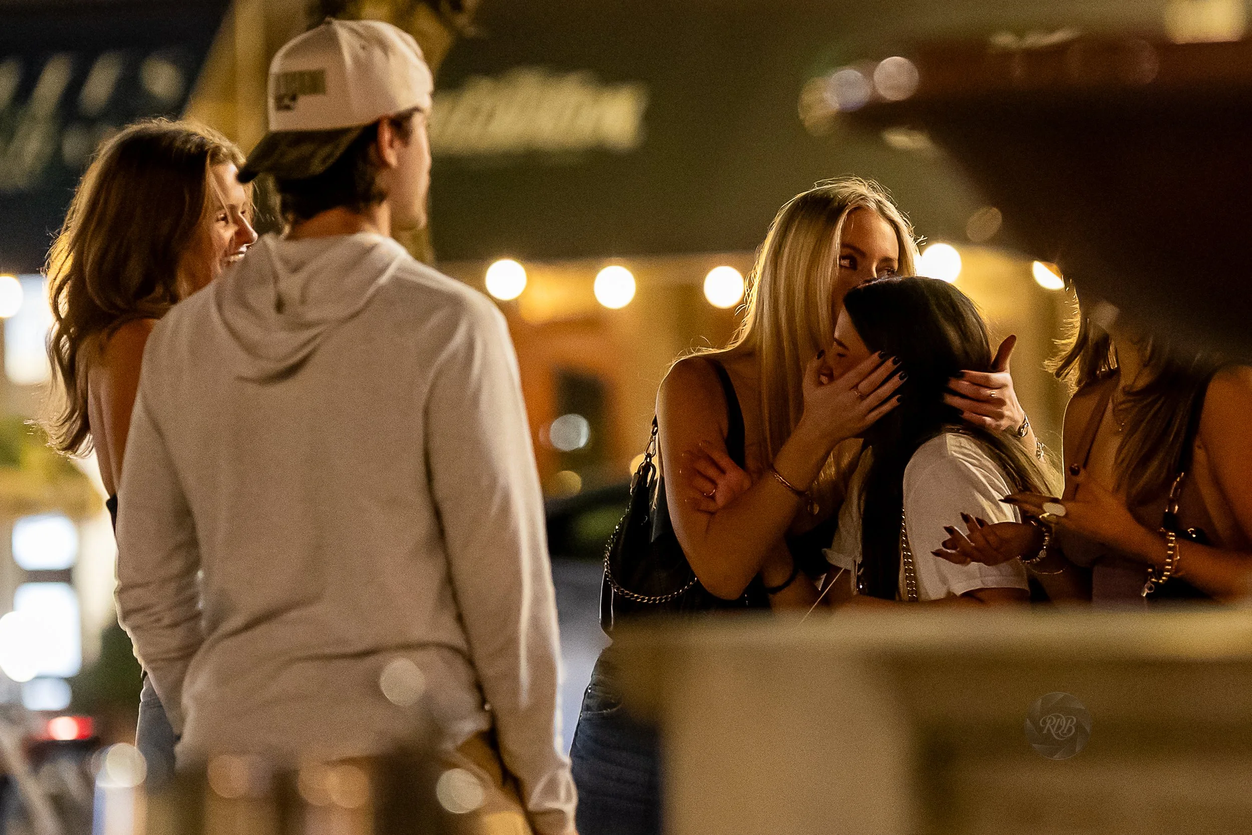 Four women and one man socializing outside at night, with string lights and a blurred city background.