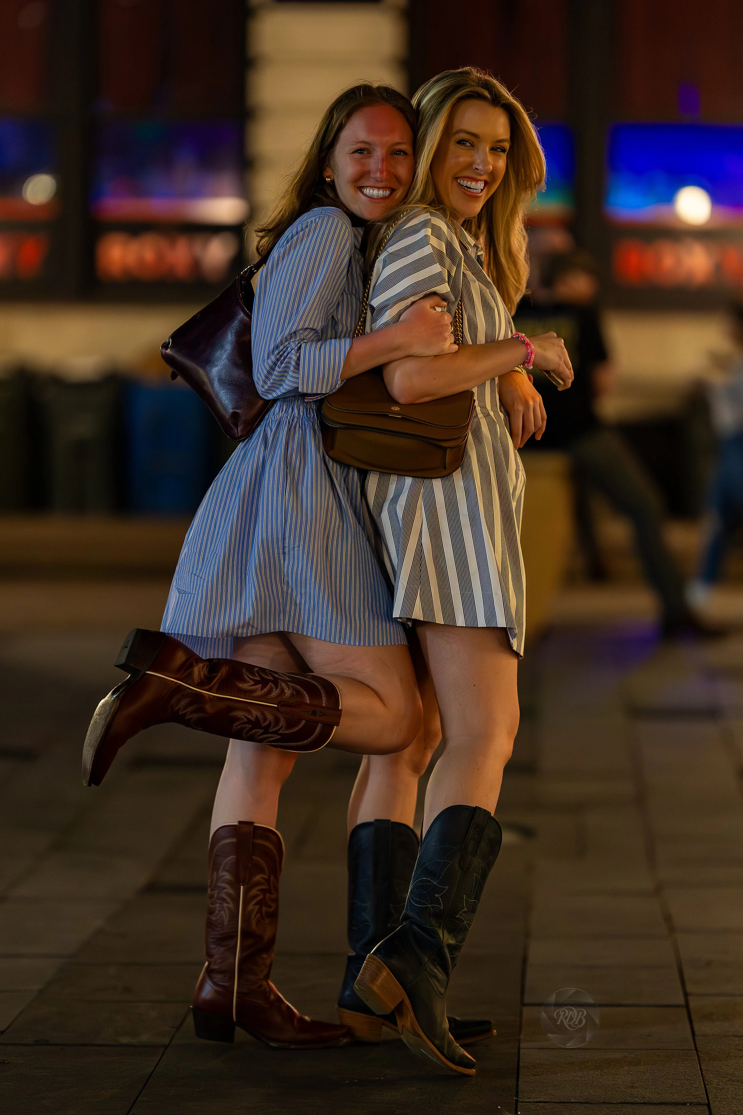 Two women smiling and hugging each other, dressed in striped dresses, wearing cowboy boots, standing on a wooden floor in a lively indoor setting with blurry colorful lights in the background.
