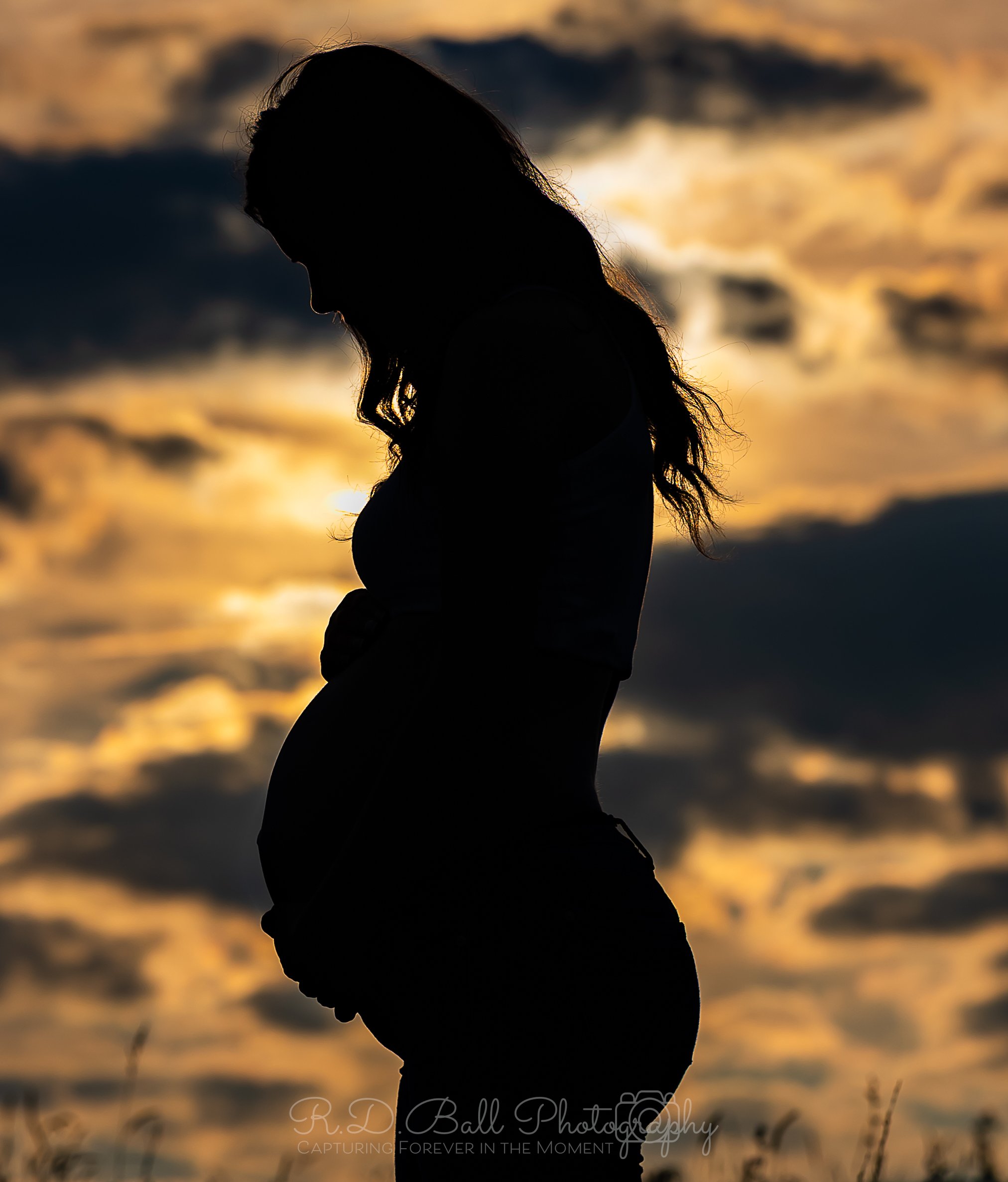 Silhouette of a pregnant woman standing outdoors during sunset, with her head bowed and one hand on her belly.