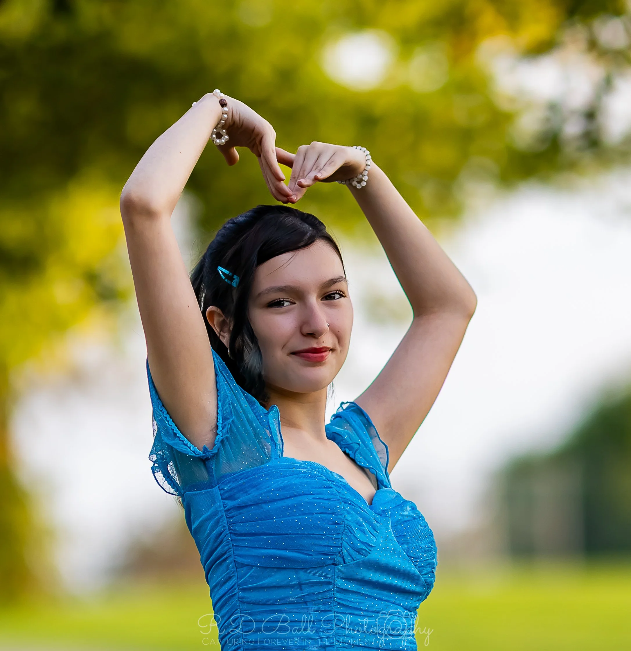 A young woman with dark hair and light skin wearing a blue dress, standing outdoors, forming a heart shape with her hands above her head, with blurred green and yellow trees in the background.