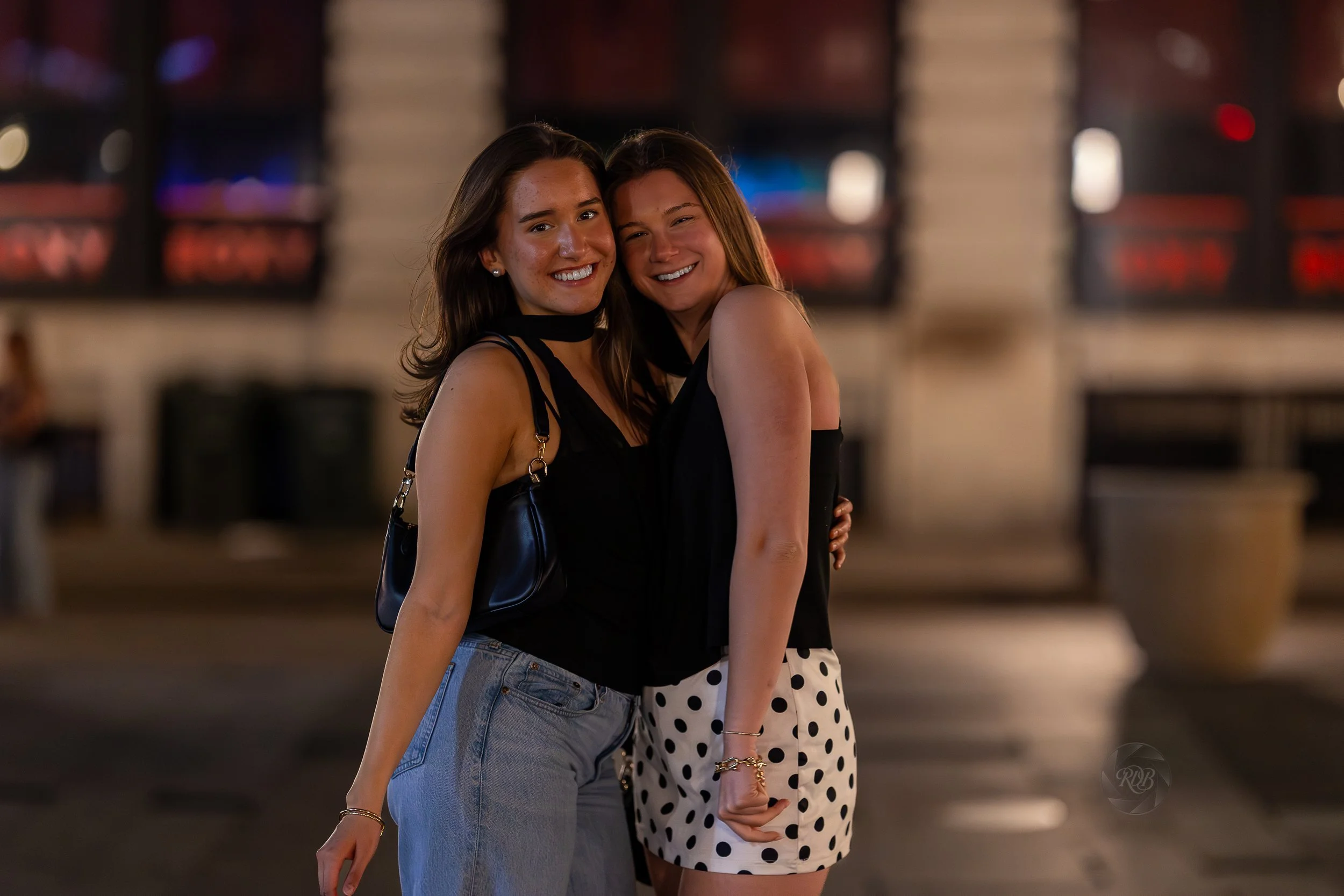 Two young women smiling and hugging each other at night in an urban setting.