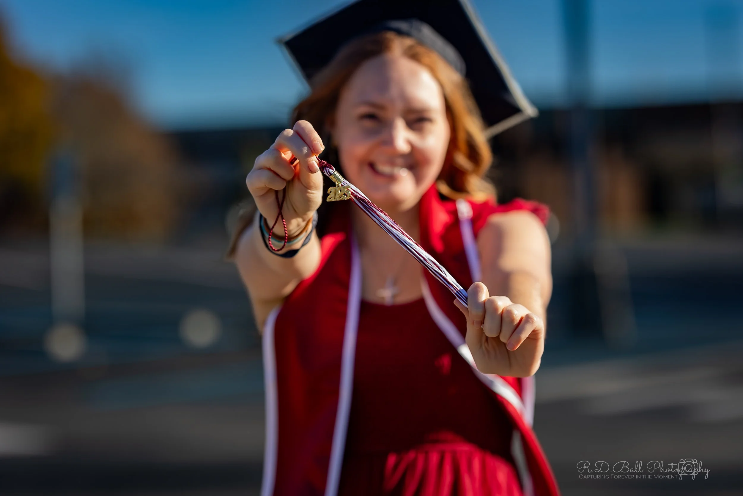 Young woman in a red dress and graduation cap smiling and holding out a 2019 graduation medal towards the camera in an outdoor setting