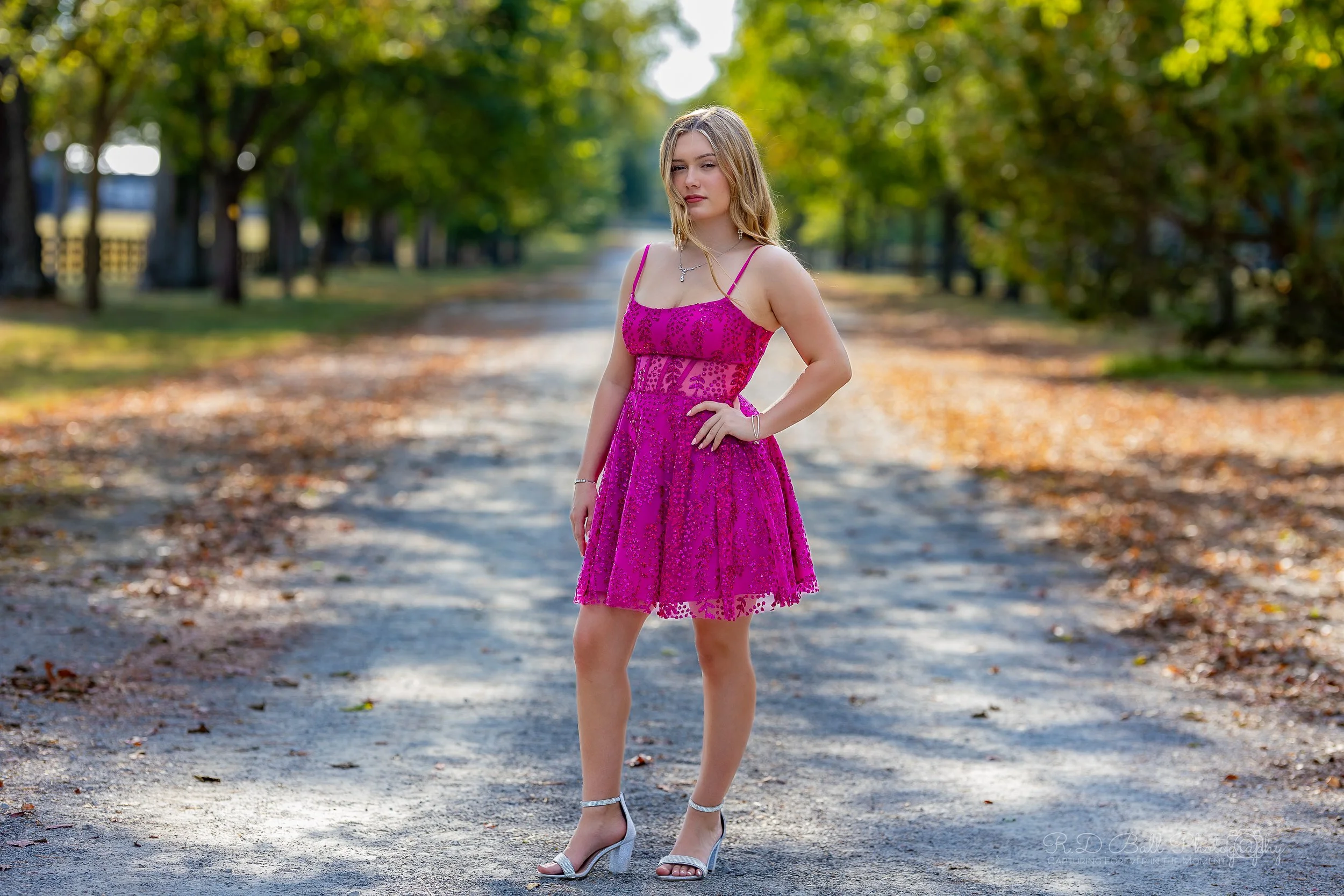A young woman in a pink lace dress and heels standing on a tree-lined dirt road with fallen leaves.