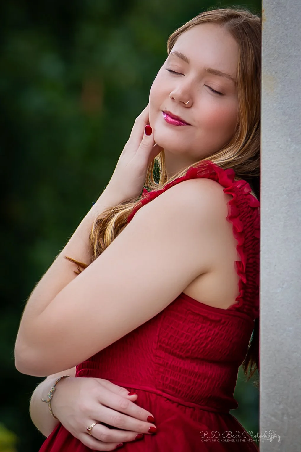 A young woman with long, wavy blond hair, closed eyes, and a gentle smile, leaning against a stone wall outdoors. She is wearing a red sleeveless dress with ruffled straps, a nose ring, red lipstick, and matching red nail polish. Her hand is resting on her chin, and she appears to be relaxed and content.