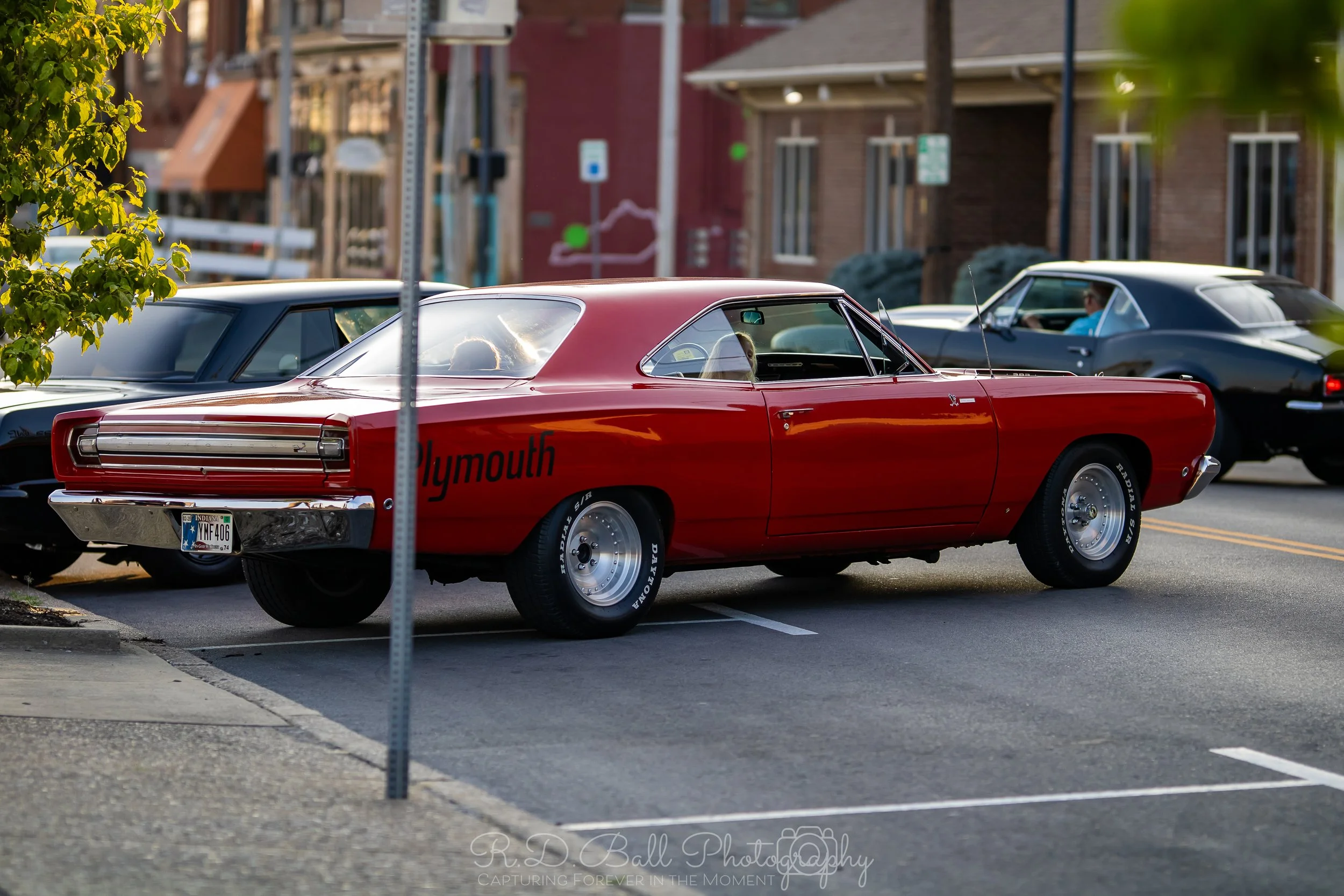 A red vintage Plymouth car parked on the street, with 'Plymouth' written on the side, surrounded by other parked cars and buildings in the background.