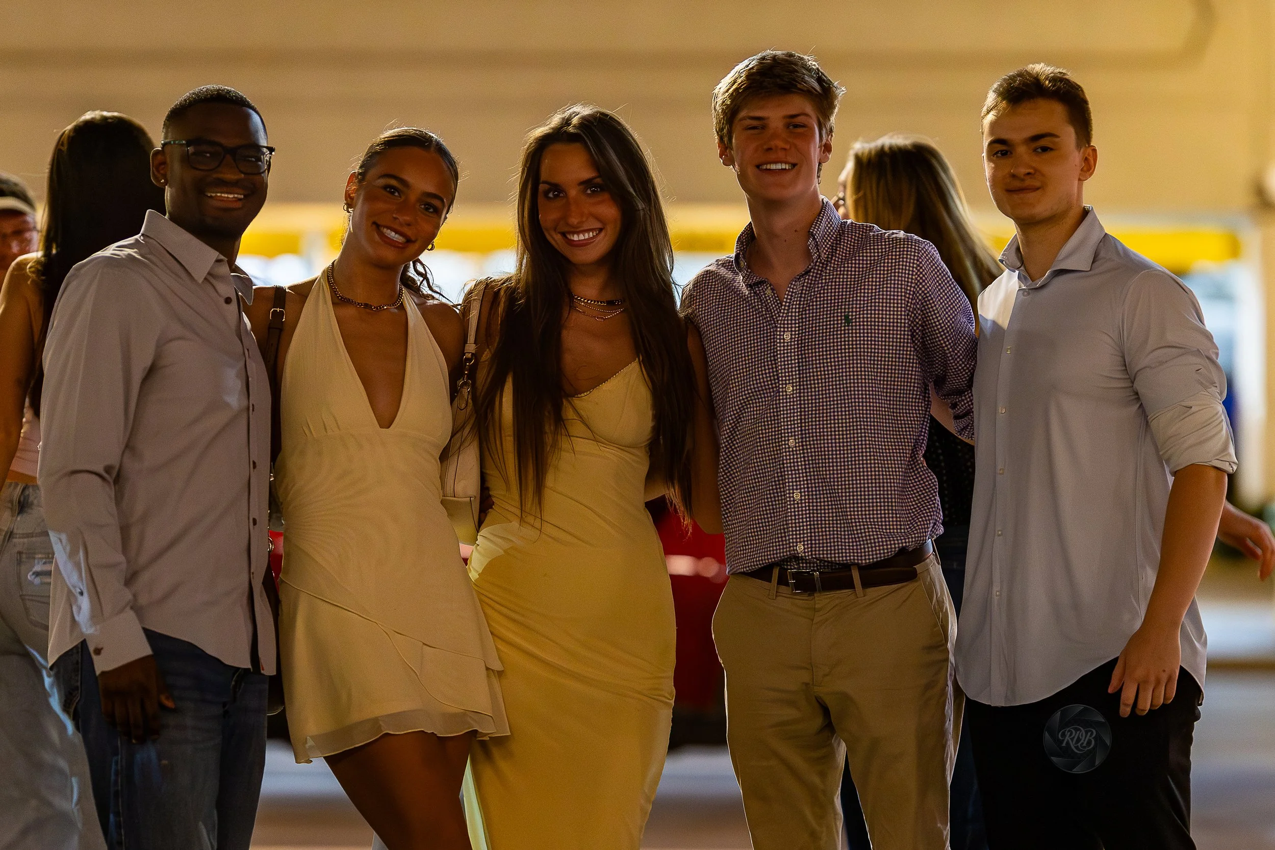 Group of five young adults smiling and standing together indoors at a social event, with a blurred background of other people and warm lighting.