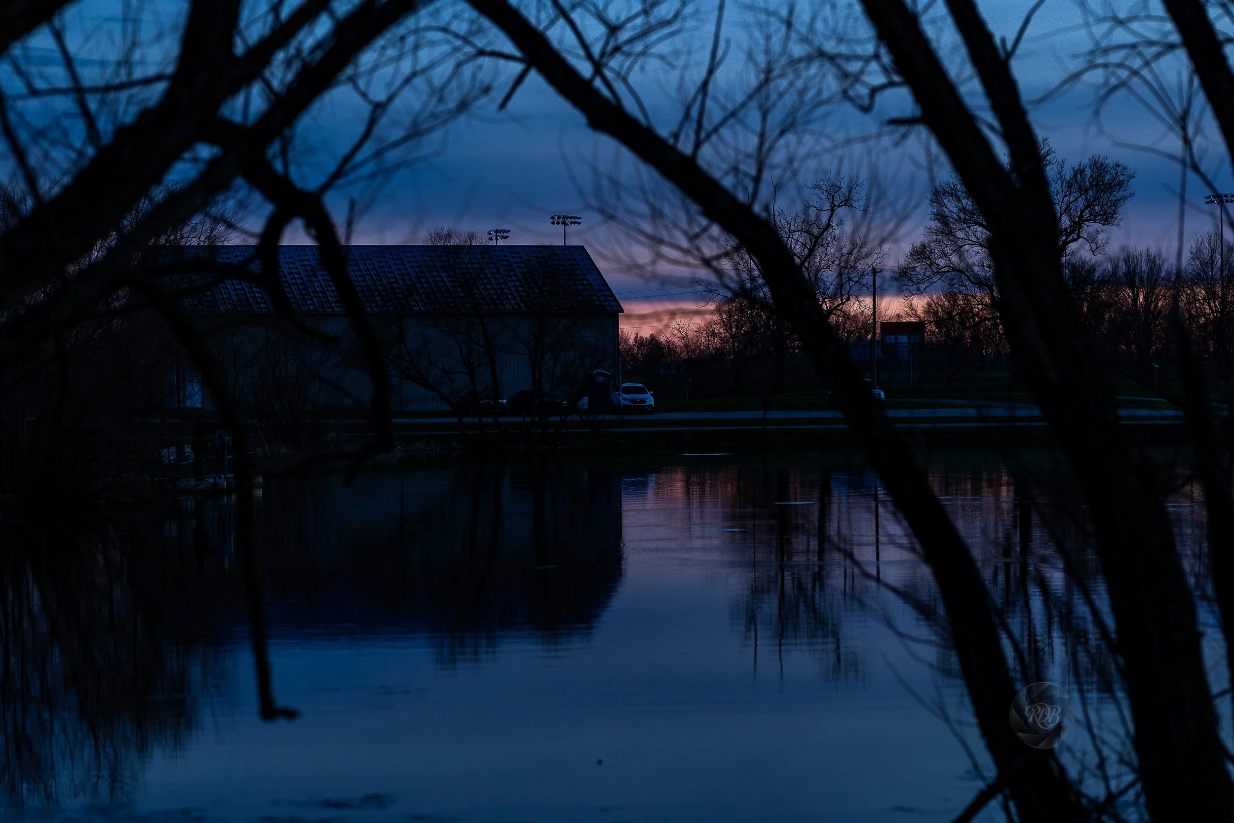 A sunset with darkening blue skies, seen through the bare branches of trees over a body of water. In the background, there is a building with a sloped roof, a few parked cars, and sports field lights.