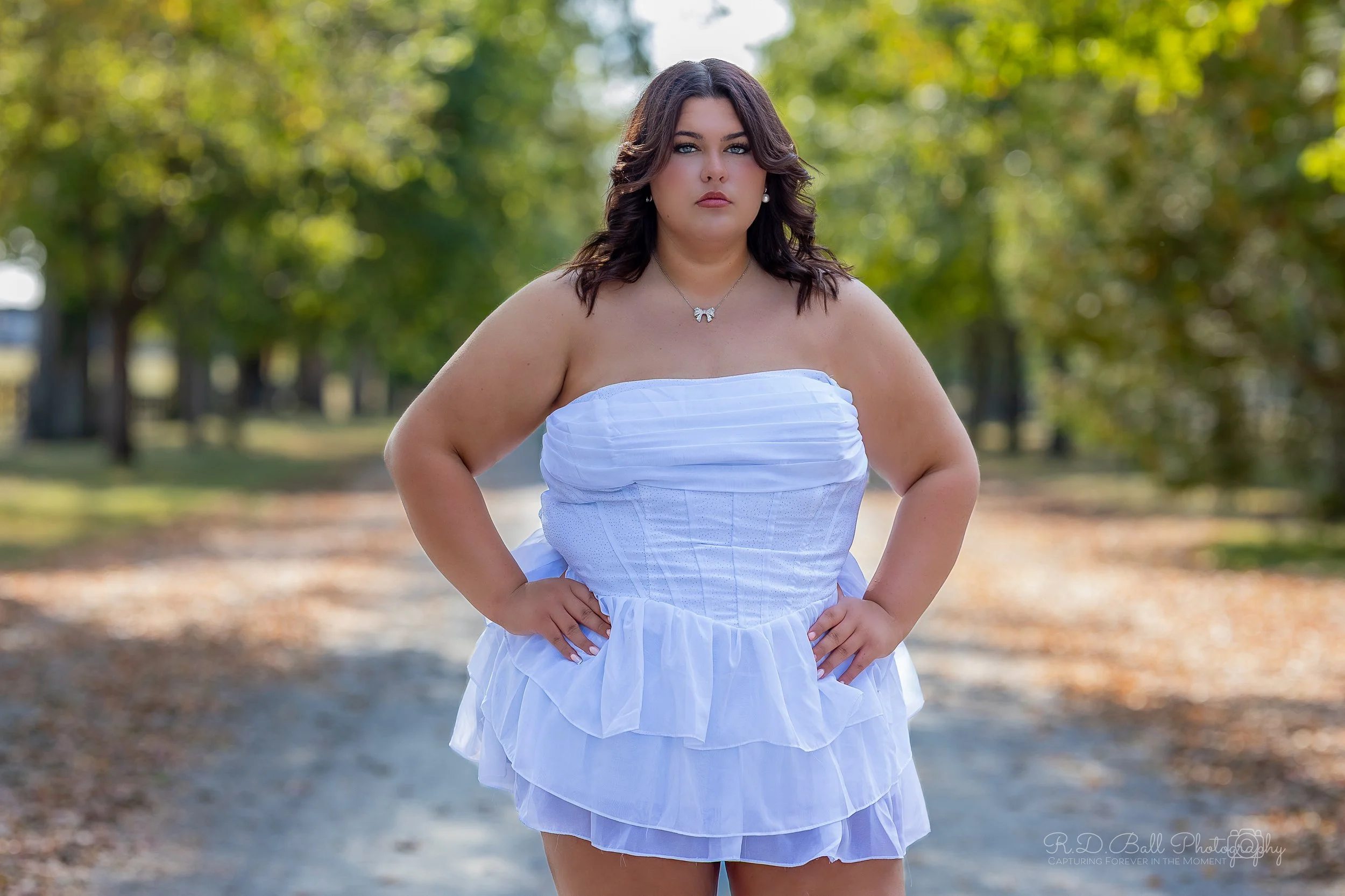 A woman standing outdoors on a dirt path surrounded by trees, wearing a strapless white dress with her hands on her hips.