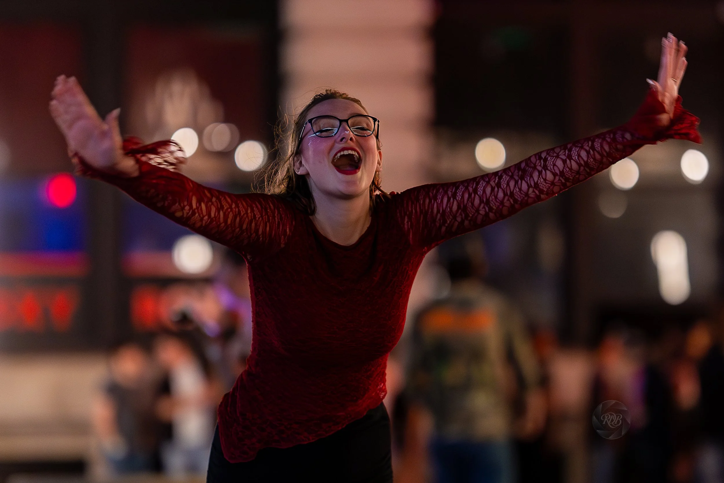 A woman with glasses and red lipstick wearing a red lace top with outstretched arms laughing in a lively, dimly lit event or party setting.
