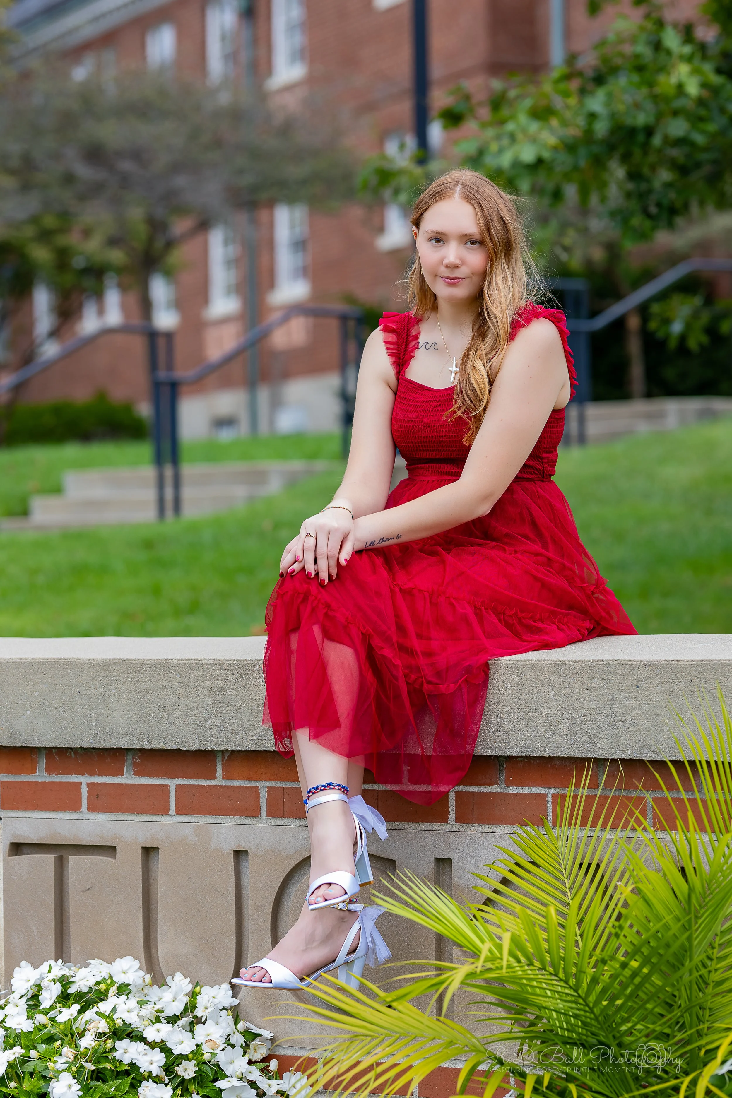 A young woman in a red dress sitting on a brick wall outdoors with green grass, trees, and a brick building in the background.