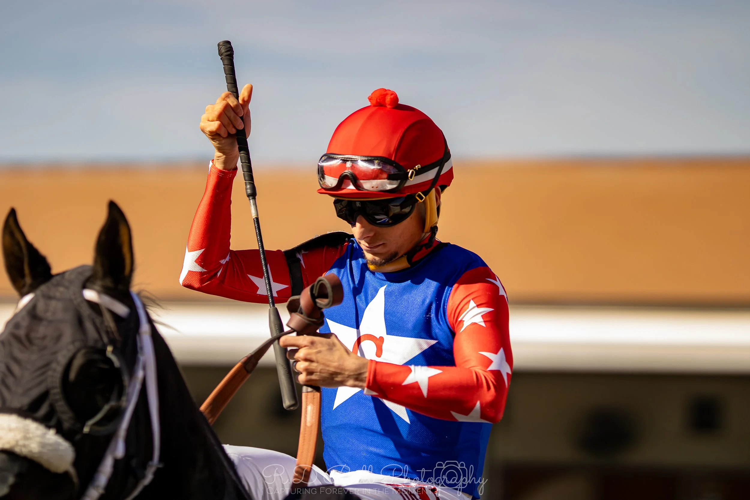 A jockey dressed in red, blue, and white with stars, wearing a red helmet and goggles, standing next to a horse.
