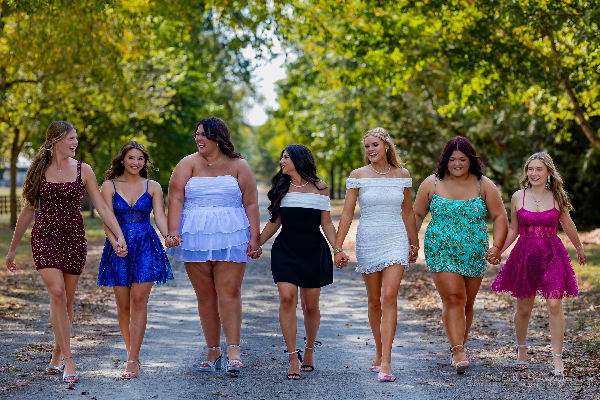 Seven women wearing colorful dresses walking hand in hand on a tree-lined path, smiling and enjoying each other's company.
