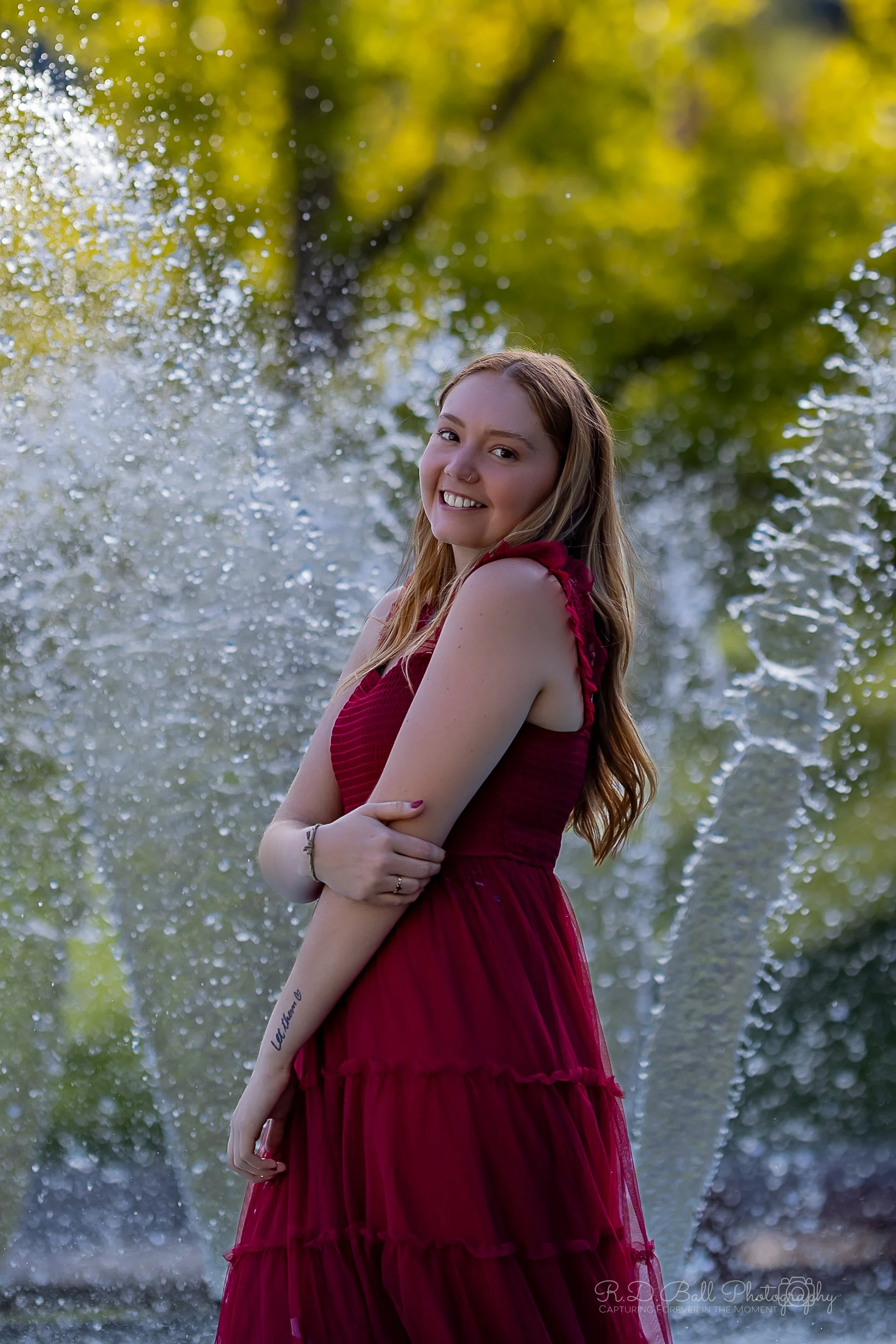 A young woman in a burgundy dress smiling and posing with arms crossed in front of a fountain with water splashing around her on a sunny day.