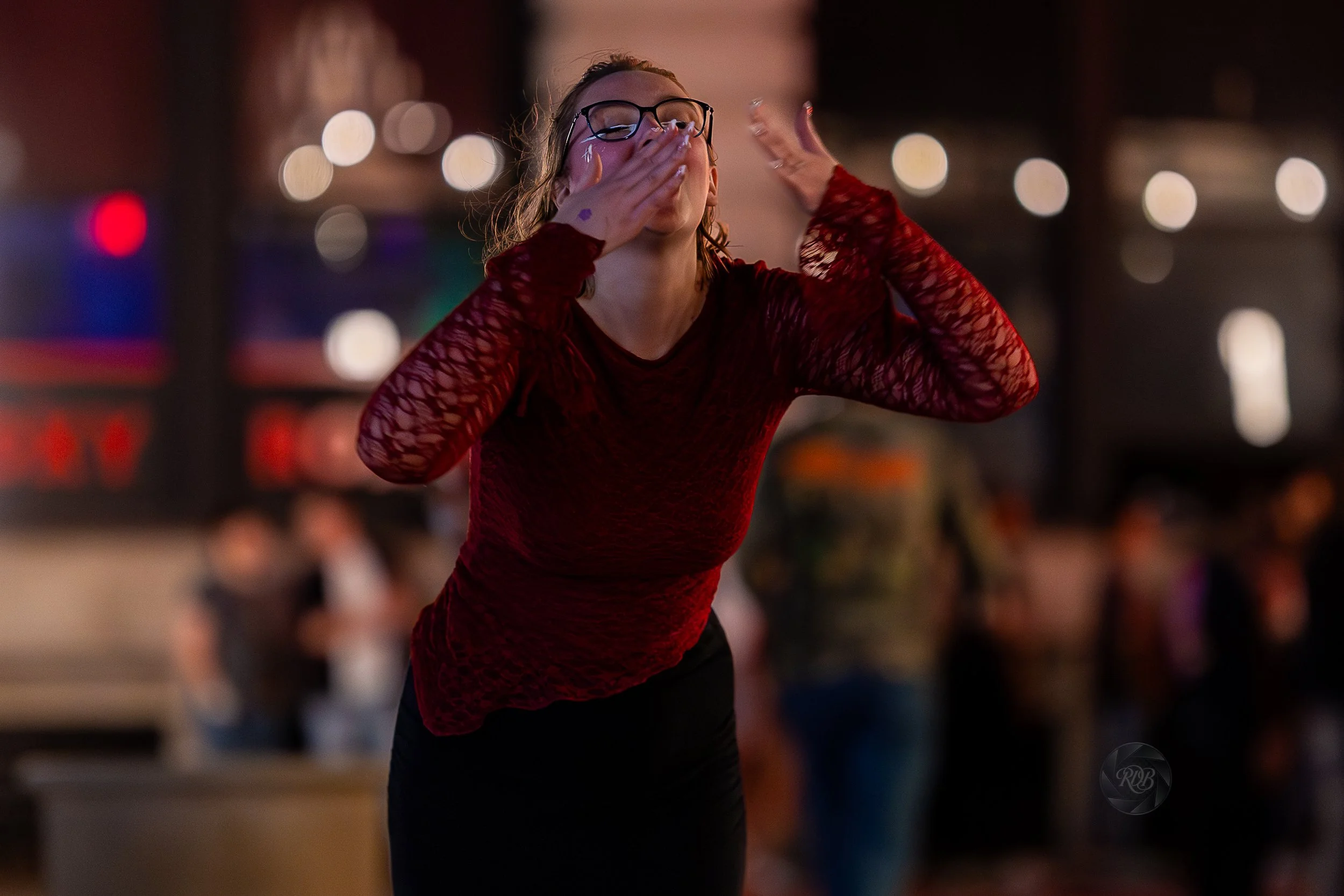 A woman in a red lace top and glasses is blowing a kiss with her hands in a lively indoor setting with blurred people in the background.