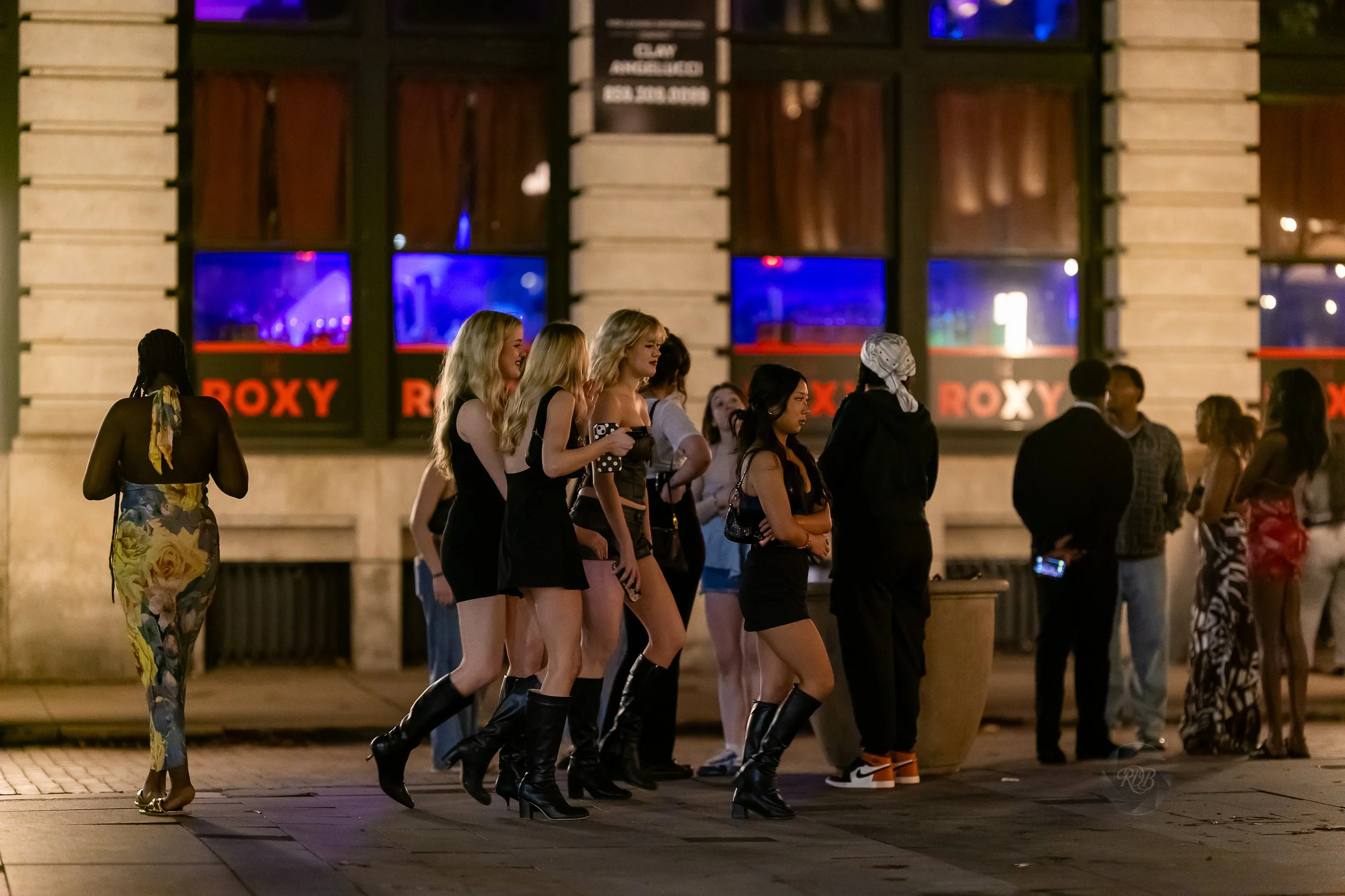 People standing and walking on a city sidewalk outside a building with illuminated windows at night.
