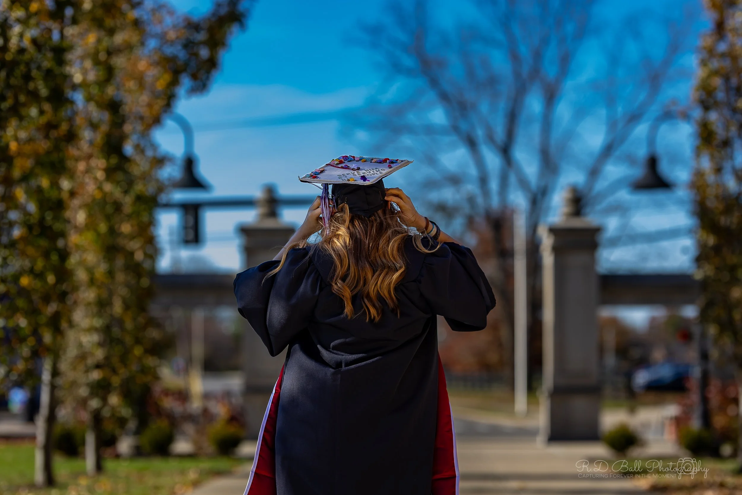 Back view of a girl in a graduation gown and cap adjusting her cap outdoors on a sunny day, with trees and a stone gate in the background.