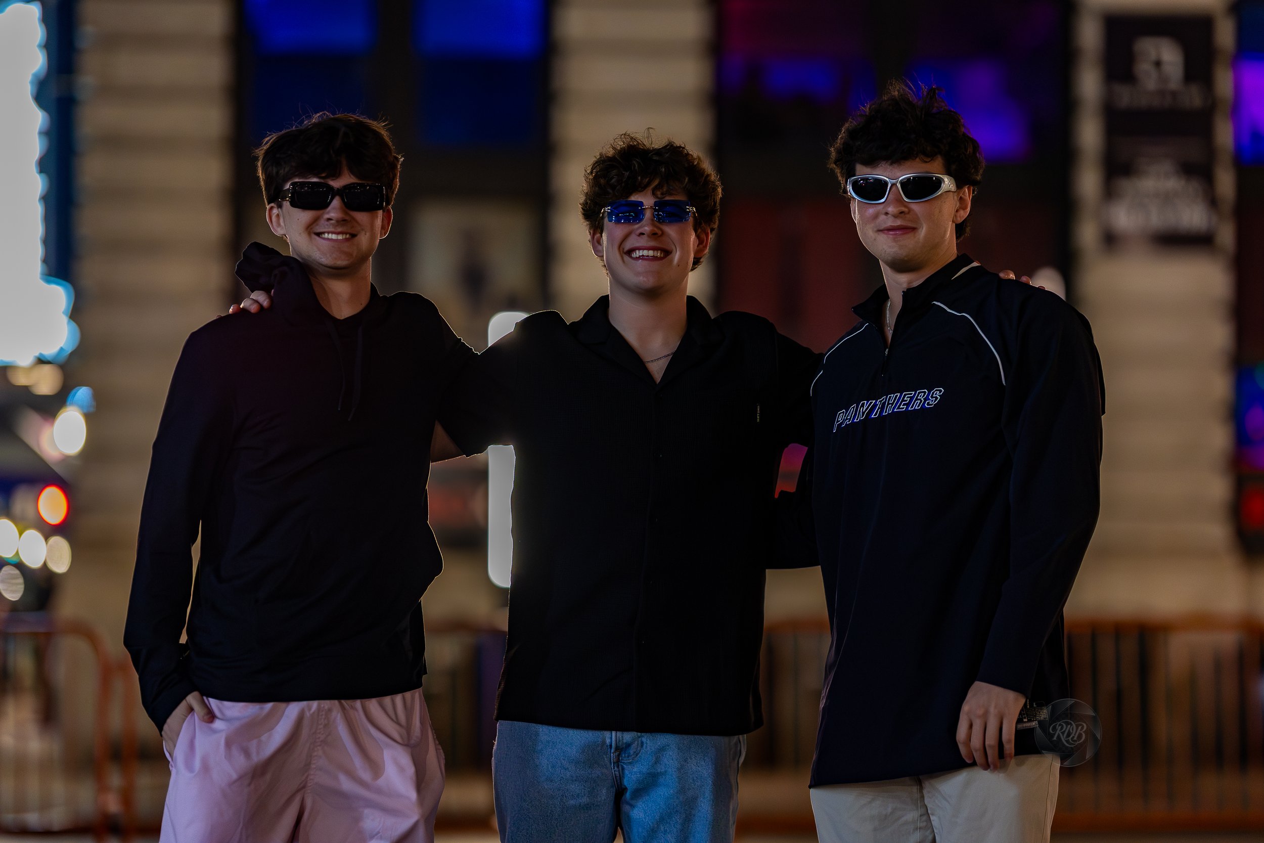 Three young men wearing sunglasses and casual clothing posing together outdoors at night, smiling.