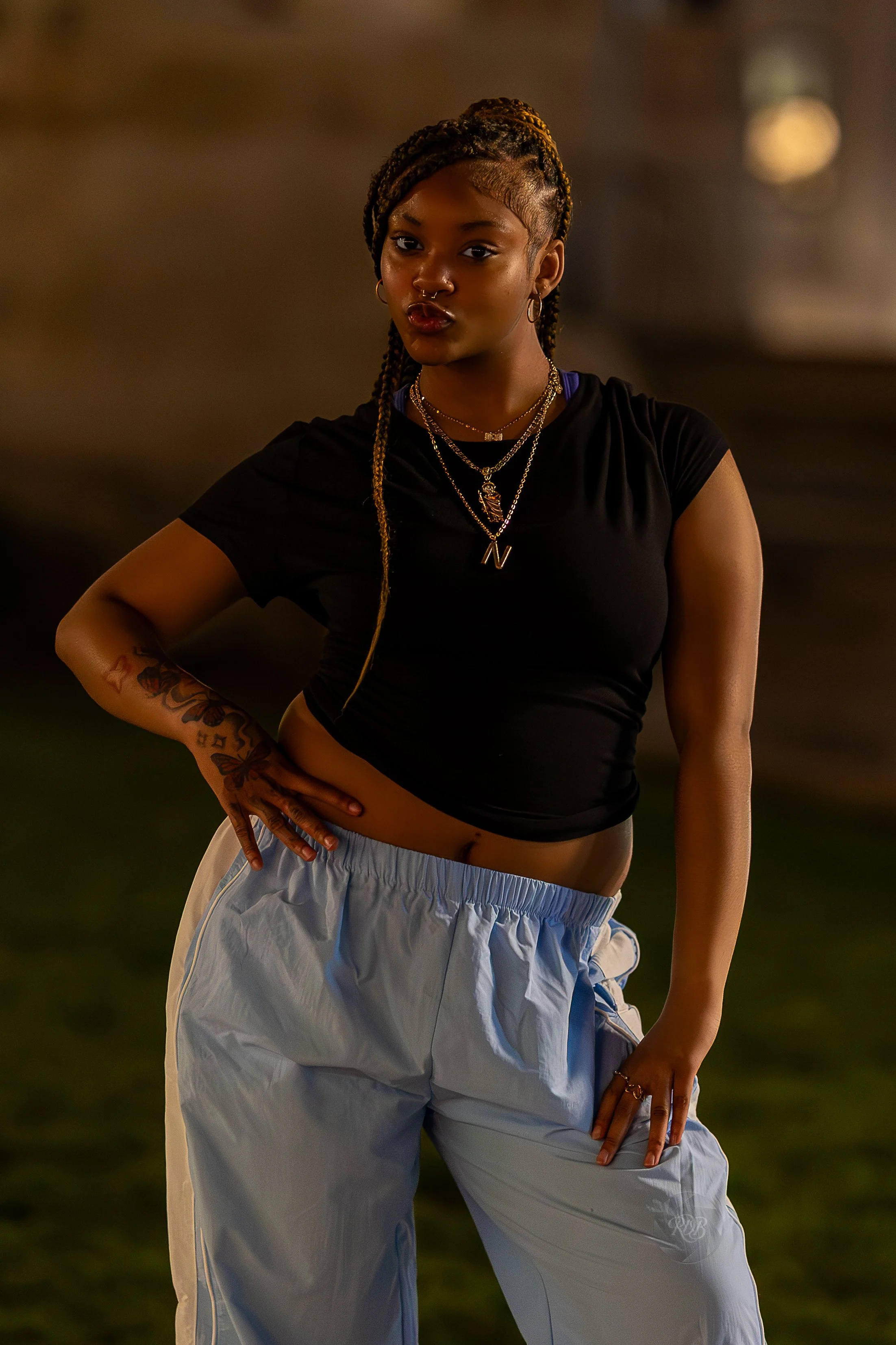 A young woman with braided hair posing with her hand on her hip in a dark setting. She's wearing a black crop top, light gray baggy pants, and layered gold necklaces.