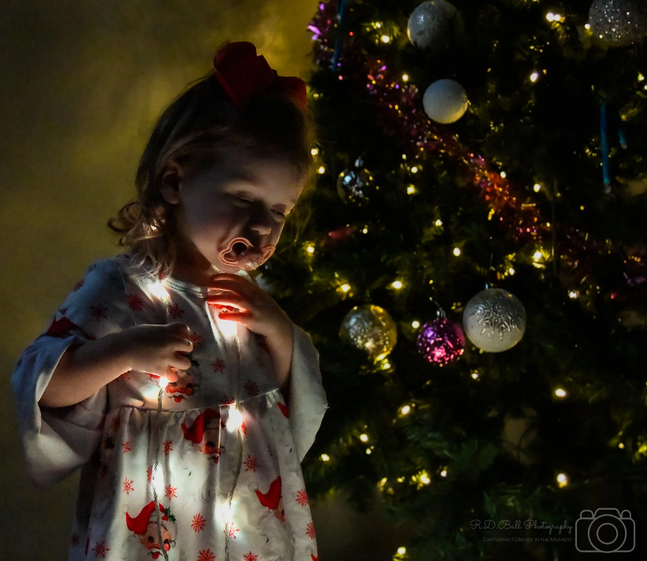 A young girl with a red bow in her hair, wearing a Christmas-themed dress with reindeer and snowflakes, stands in front of a decorated Christmas tree at night. She appears to be looking at Christmas lights with a fascinated expression, with her hands on her chest and the tree adorned with ornaments and string lights illuminating the scene.