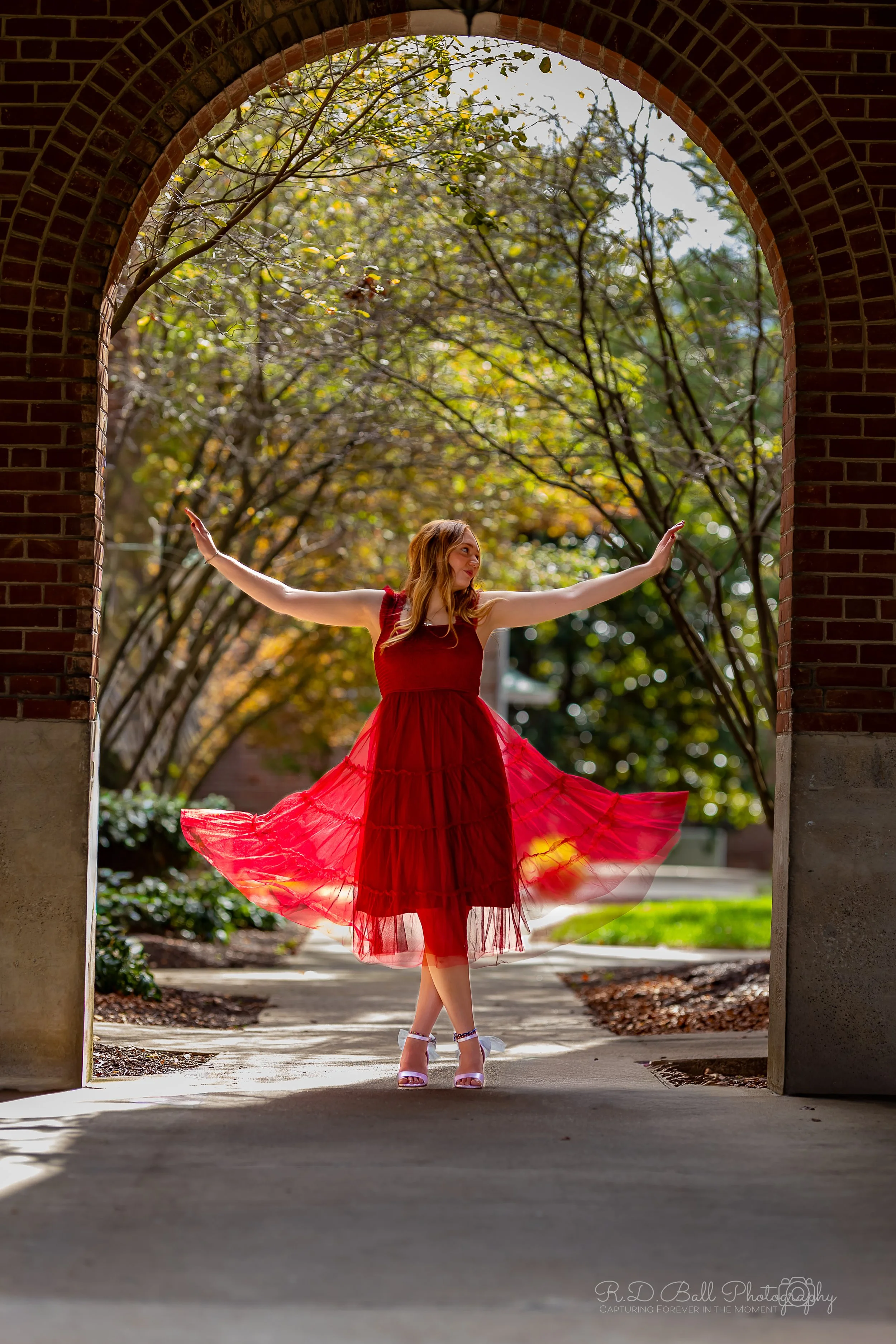 A woman in a red dress twirling under an archway with autumn trees in the background.