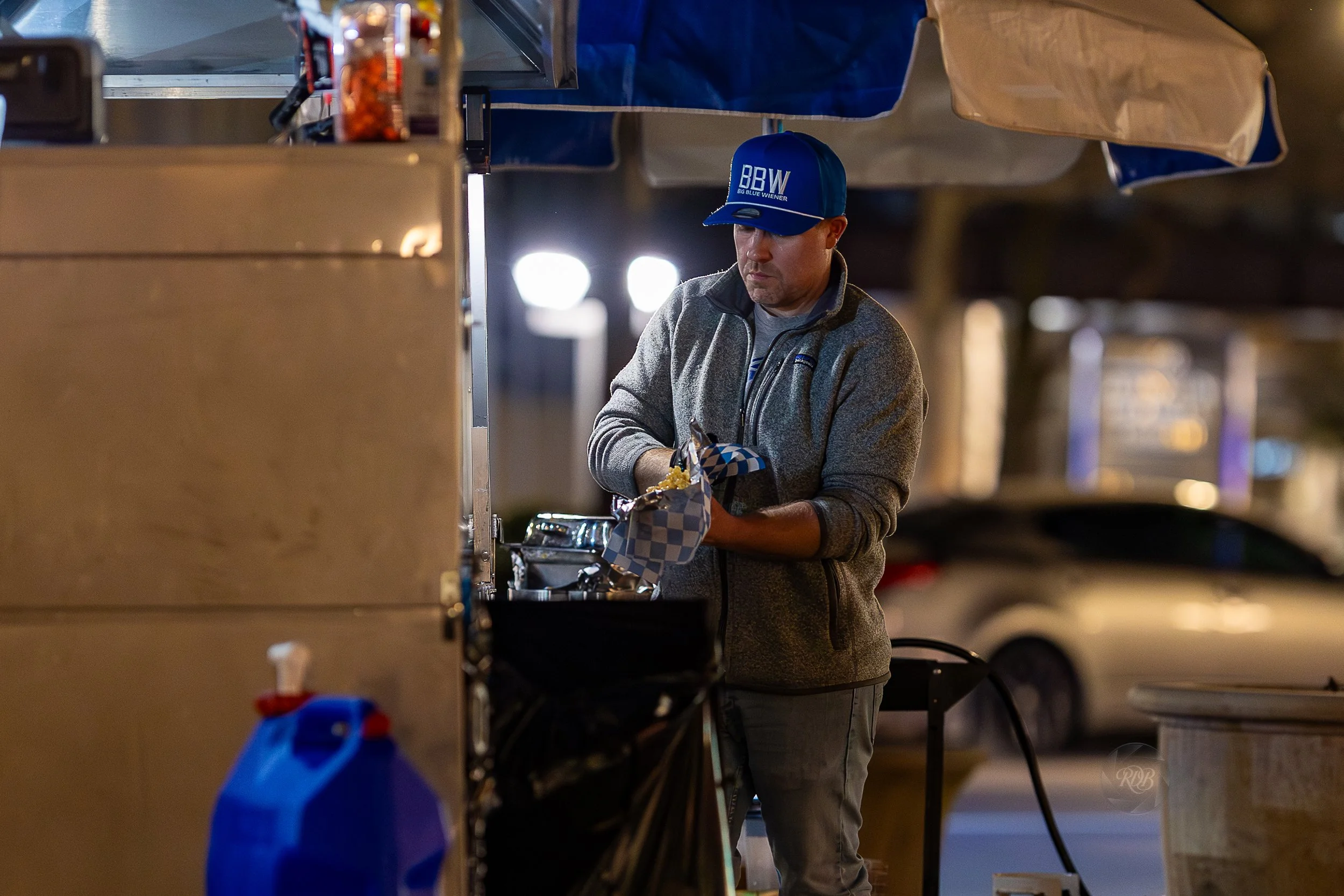 A man wearing a blue cap and gray fleece jacket preparing food at a street food cart at night.
