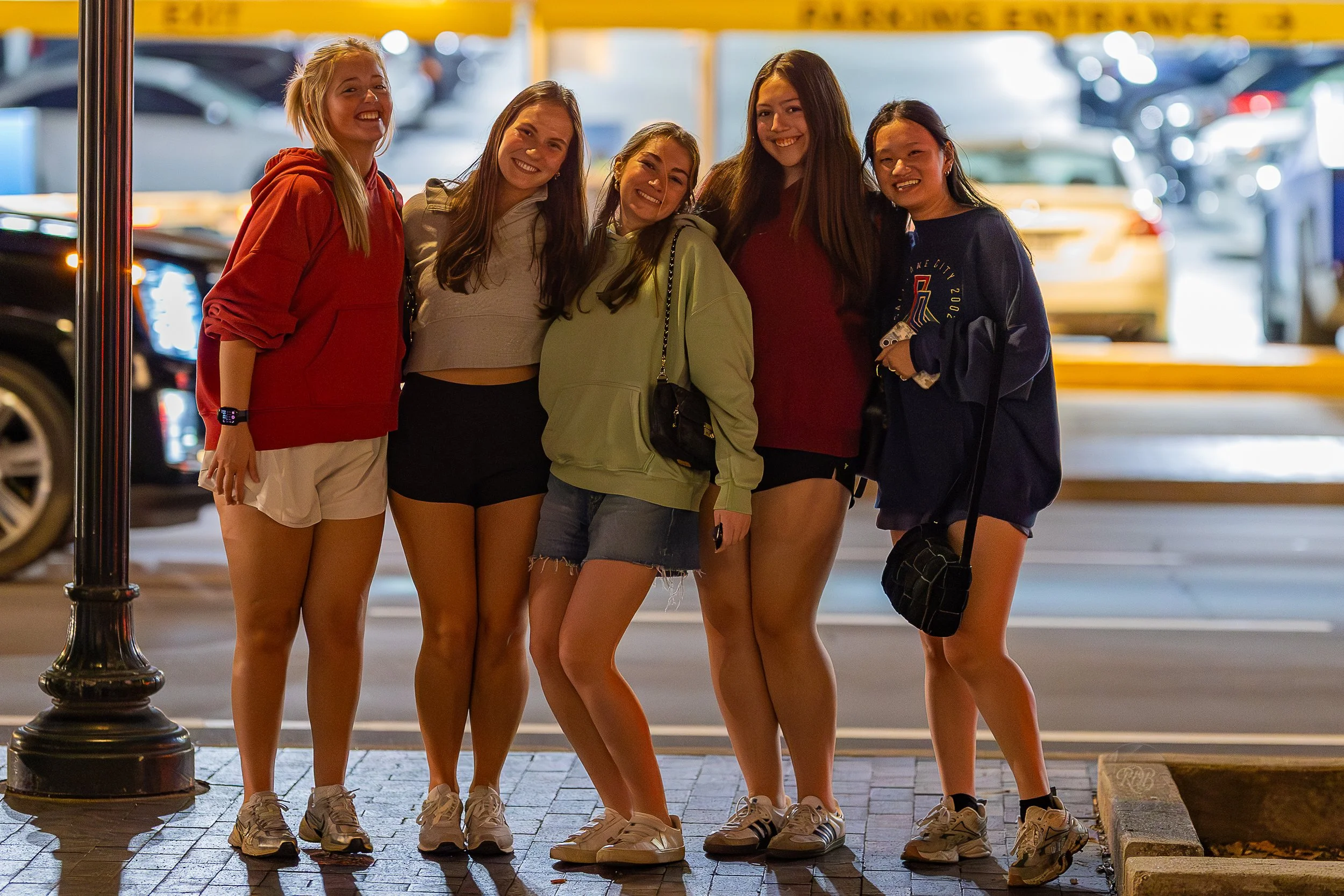 Six young women standing together on a city sidewalk at night, smiling and posing for the photo, with parked cars and yellow parking garage signage in the background.