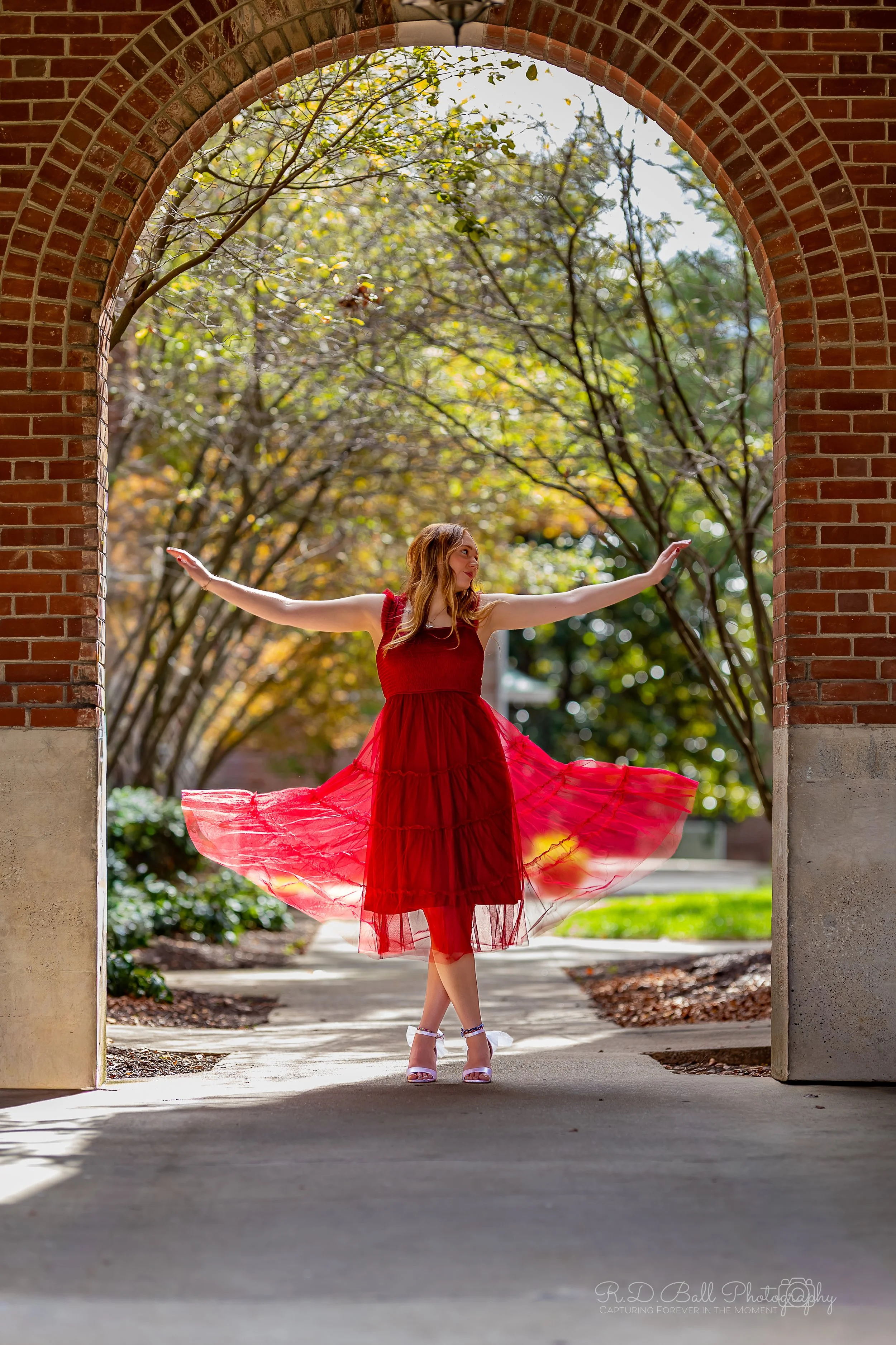 A woman in a red dress twirling under an archway surrounded by trees with autumn leaves.