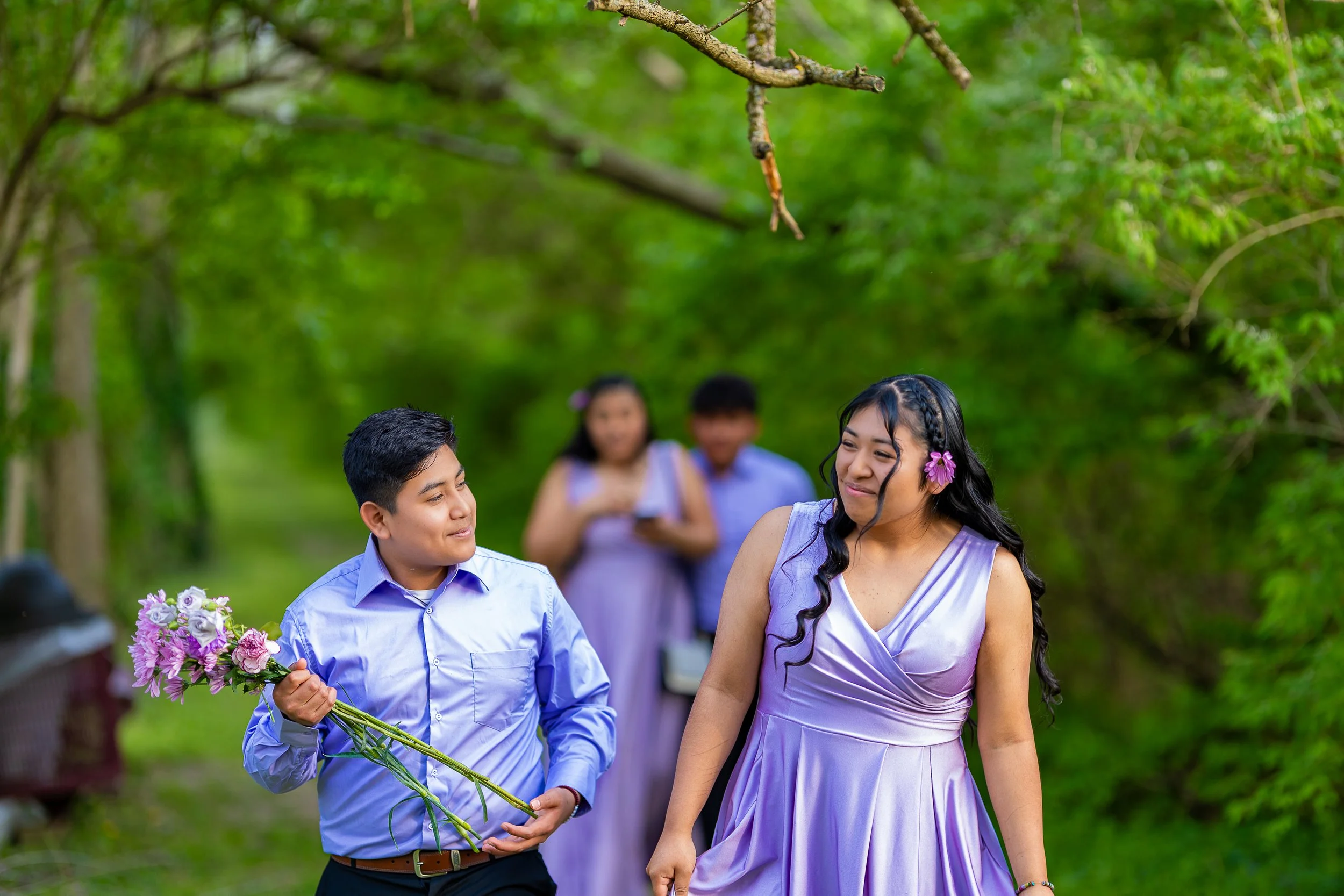 A young man in a light blue shirt holding a bouquet of pink and purple flowers, smiling at a girl in a lavender dress with a flower in her hair, in a green outdoor setting with trees, while two people watch in the background.