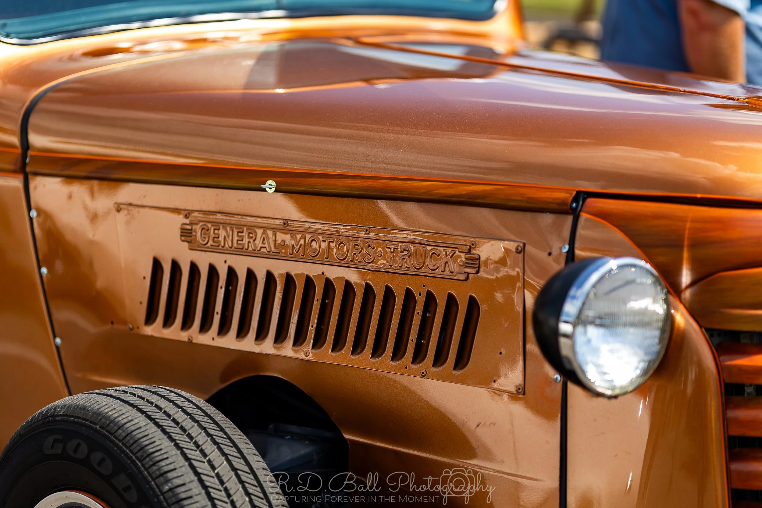 Close-up of a vintage orange truck with a vented panel on the side and a round headlight, in a rural setting.