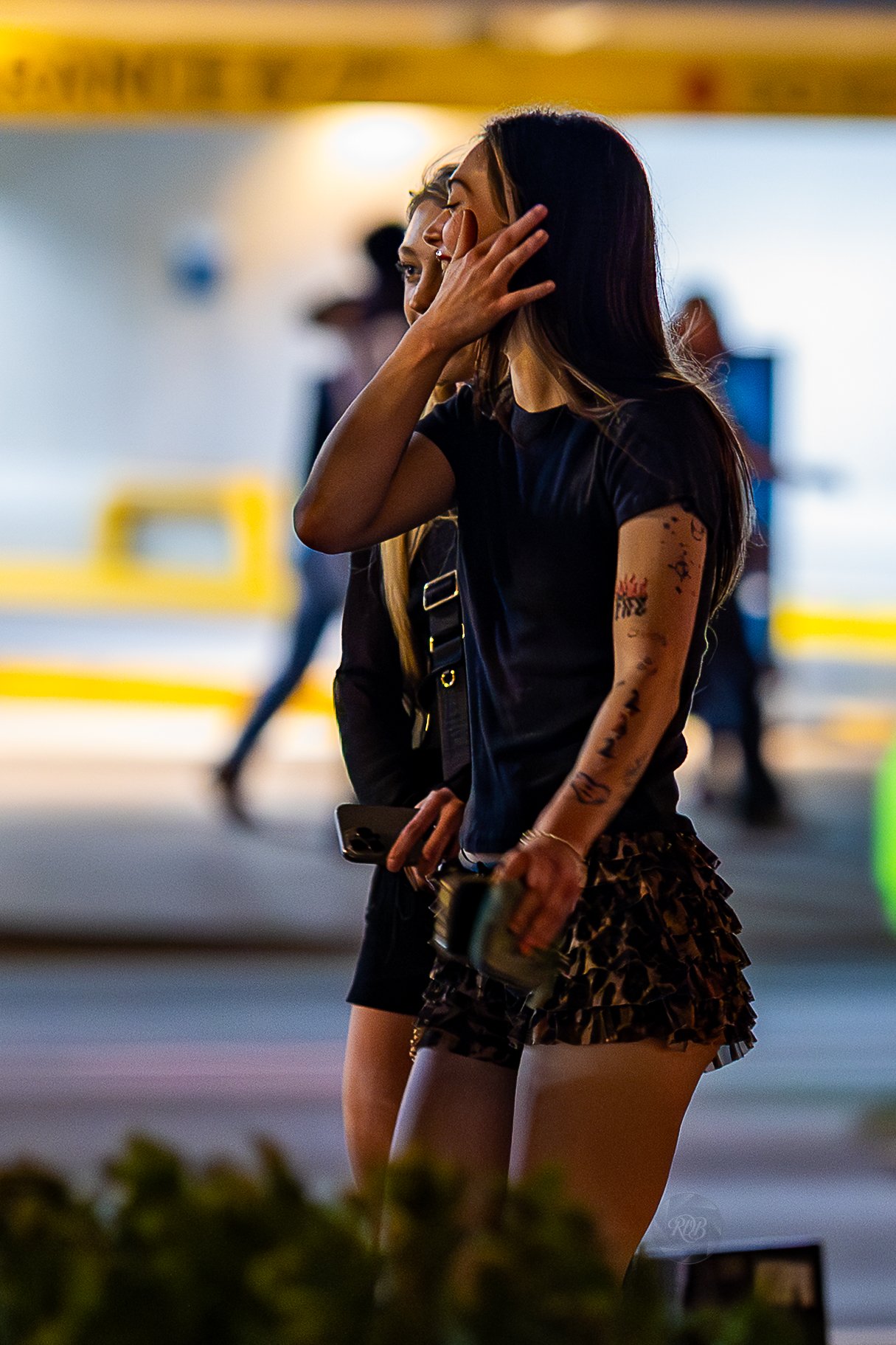 A young woman with long dark hair and tattoos on her arm, wearing a black t-shirt and animal print shorts, is standing outdoors in a city at night. She is touching her face with one hand and holding a drink and a phone in the other.