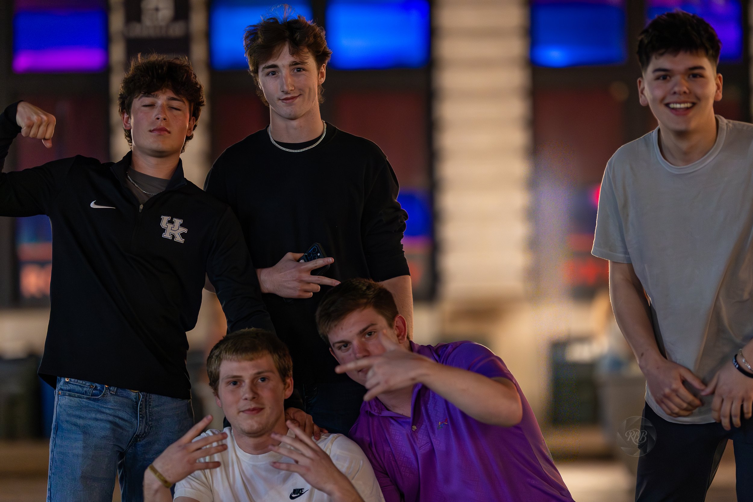 Five teenage boys posing together outdoors at night, some making gestures, with colorful lights in the background.