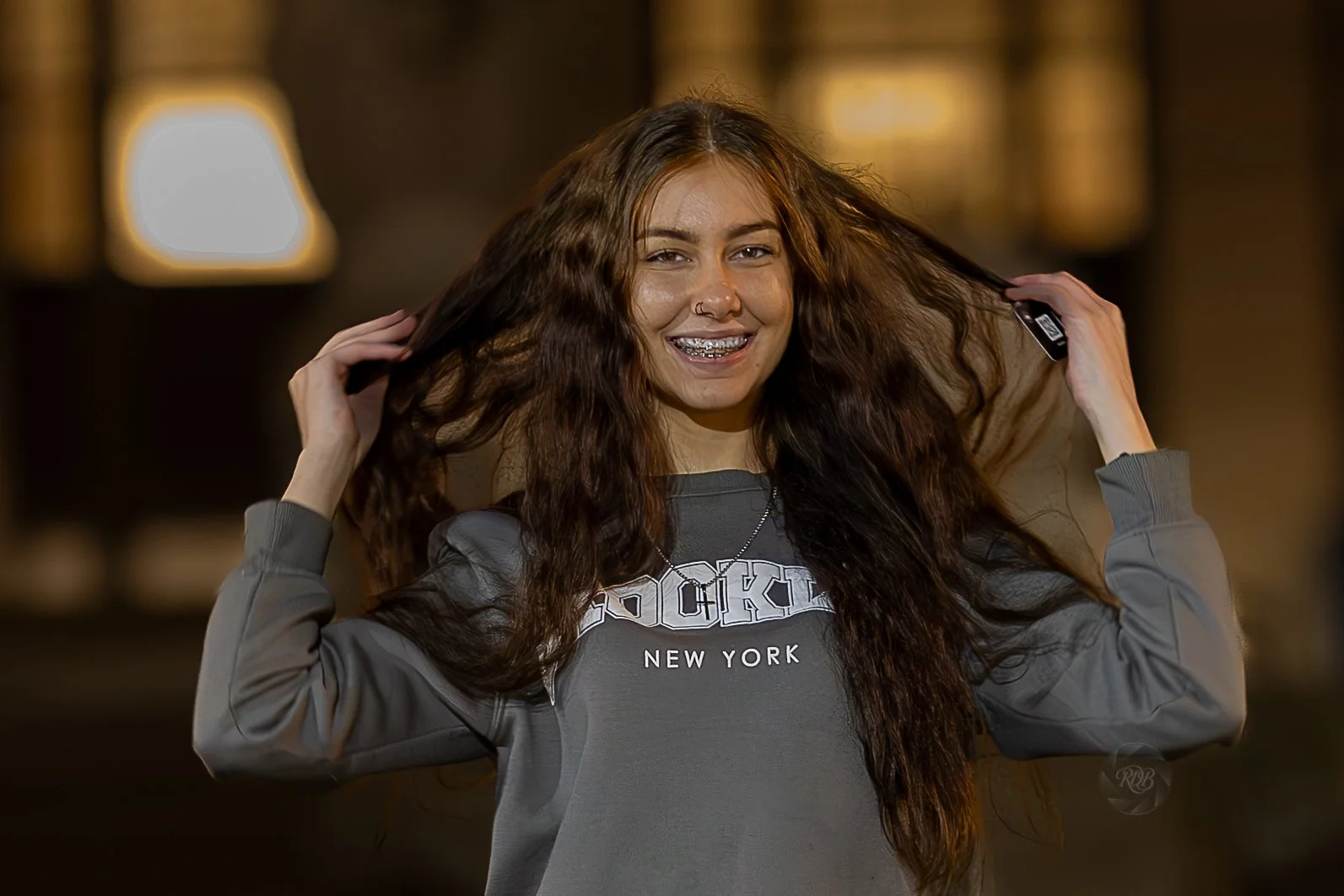 A young woman with long, curly brown hair, smiling, with braces, holding her hair with both hands, wearing a gray sweatshirt that says "ROCKY NEW YORK" in white letters, standing indoors in front of warm tone lights.