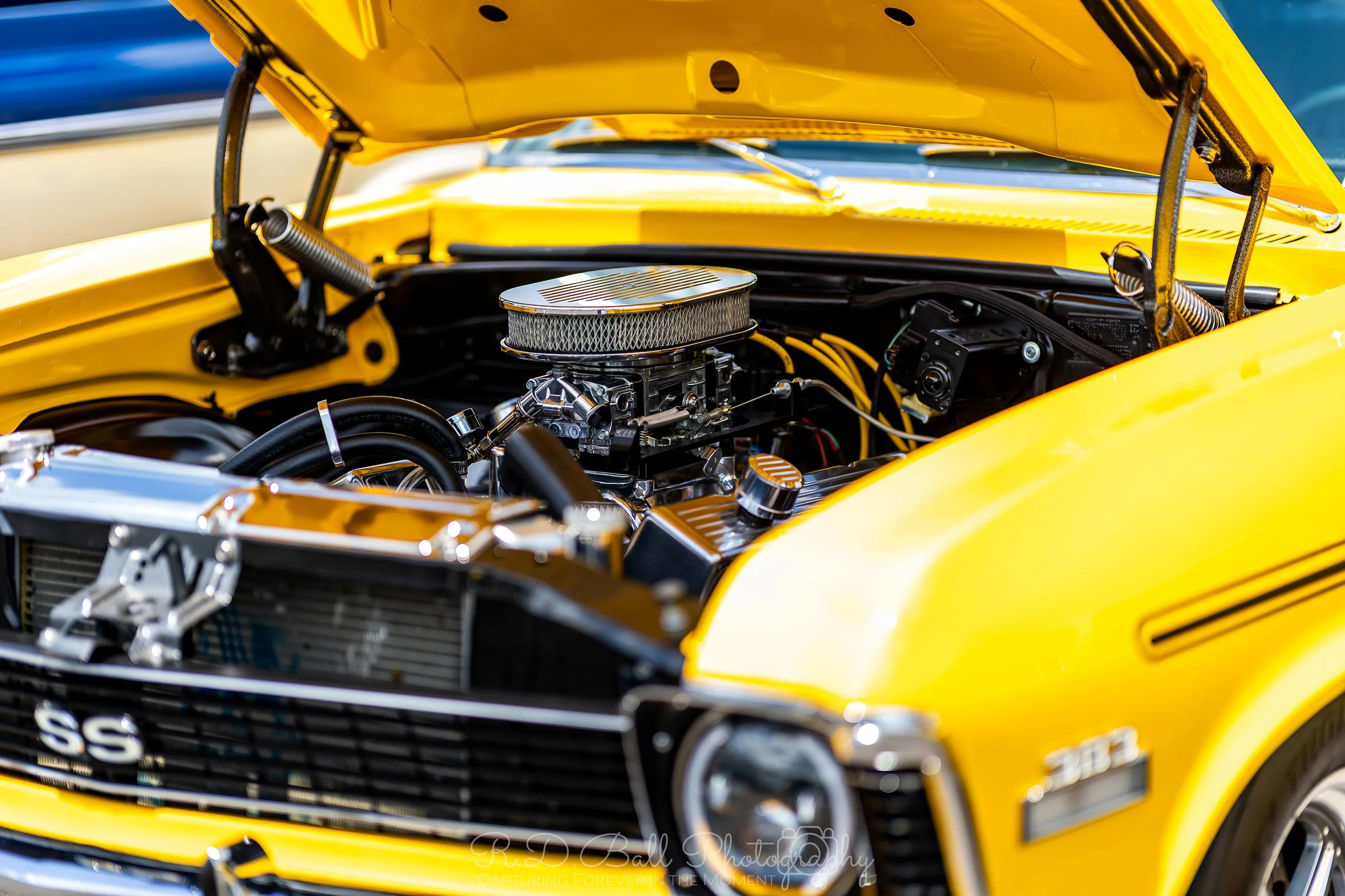 Close-up of a yellow muscle car with hood open, revealing a clean, polished engine with chrome and black components.