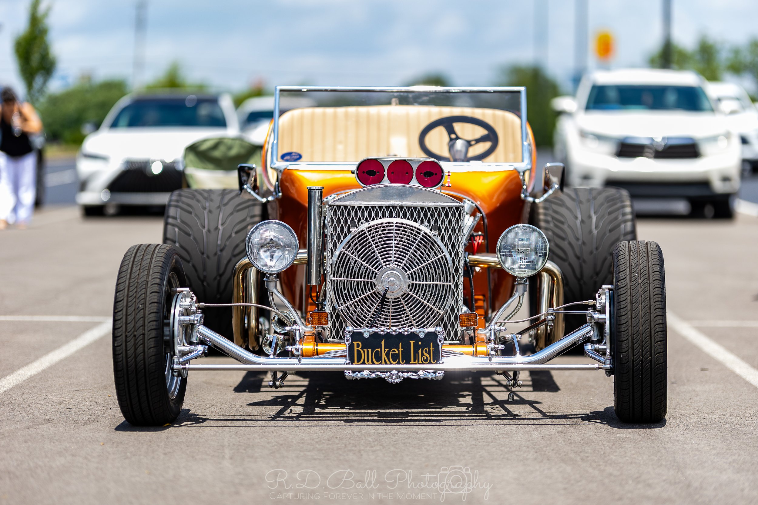 Vintage open-top race car with a large front grill and three red circles on the hood, parked in a lot with other modern cars in the background.