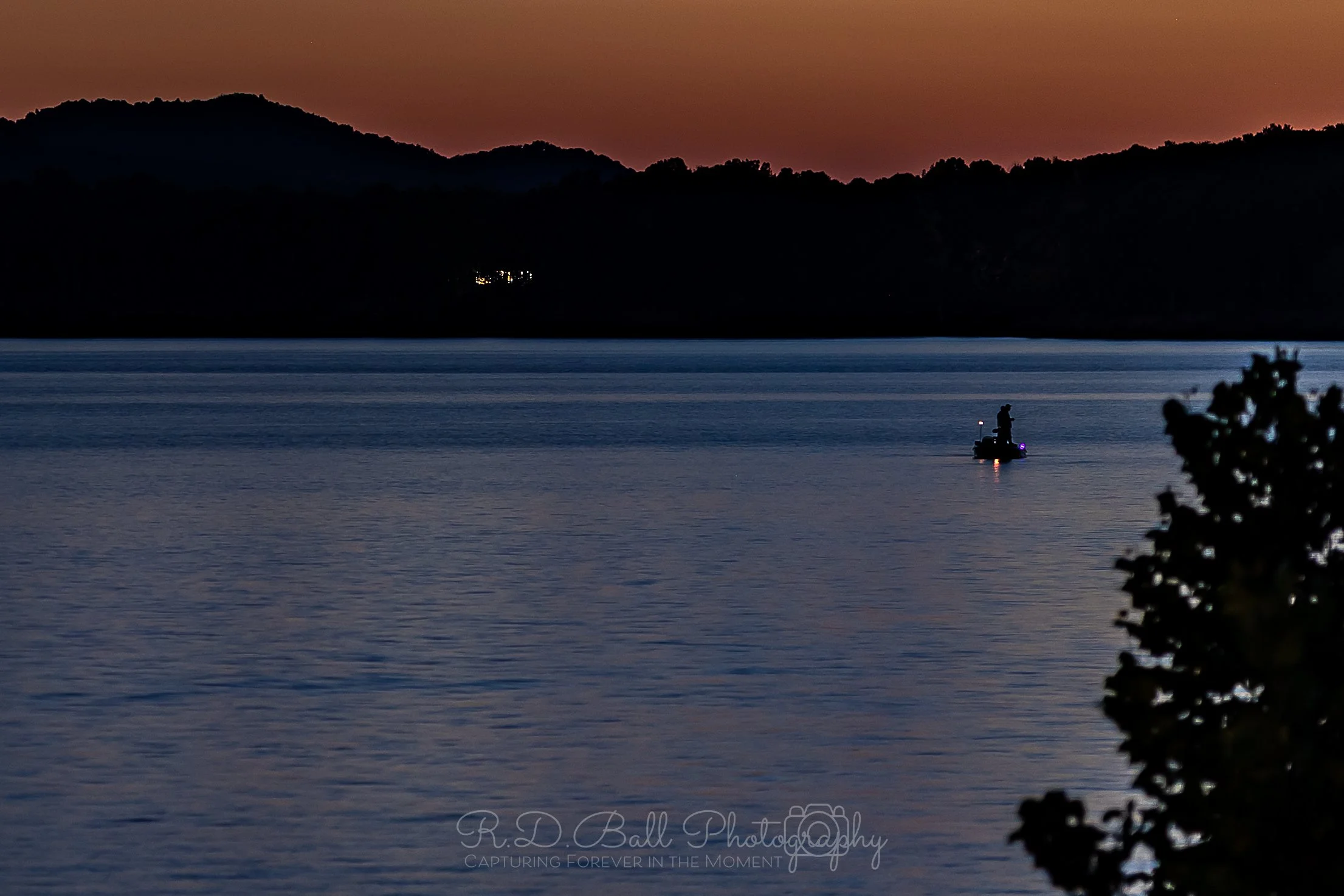 A person fishing from a boat on a calm lake at sunset, with silhouetted mountains and trees in the background.
