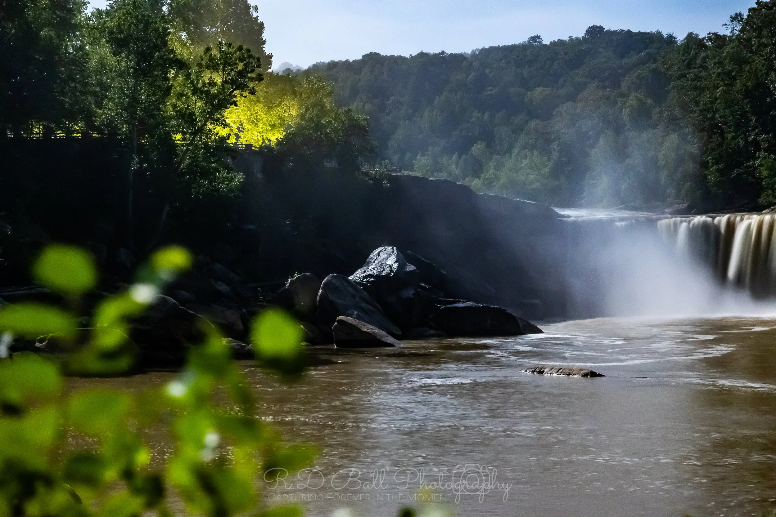 Tall waterfall flowing into a river with large rocks in the foreground, surrounded by lush green trees and forested hills in the background.