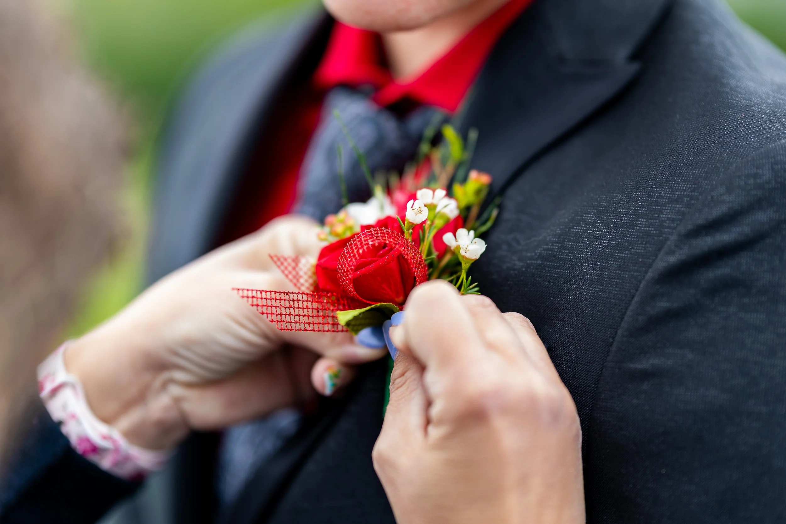 Person pinning a red and white boutonniere onto a man's dark suit jacket.