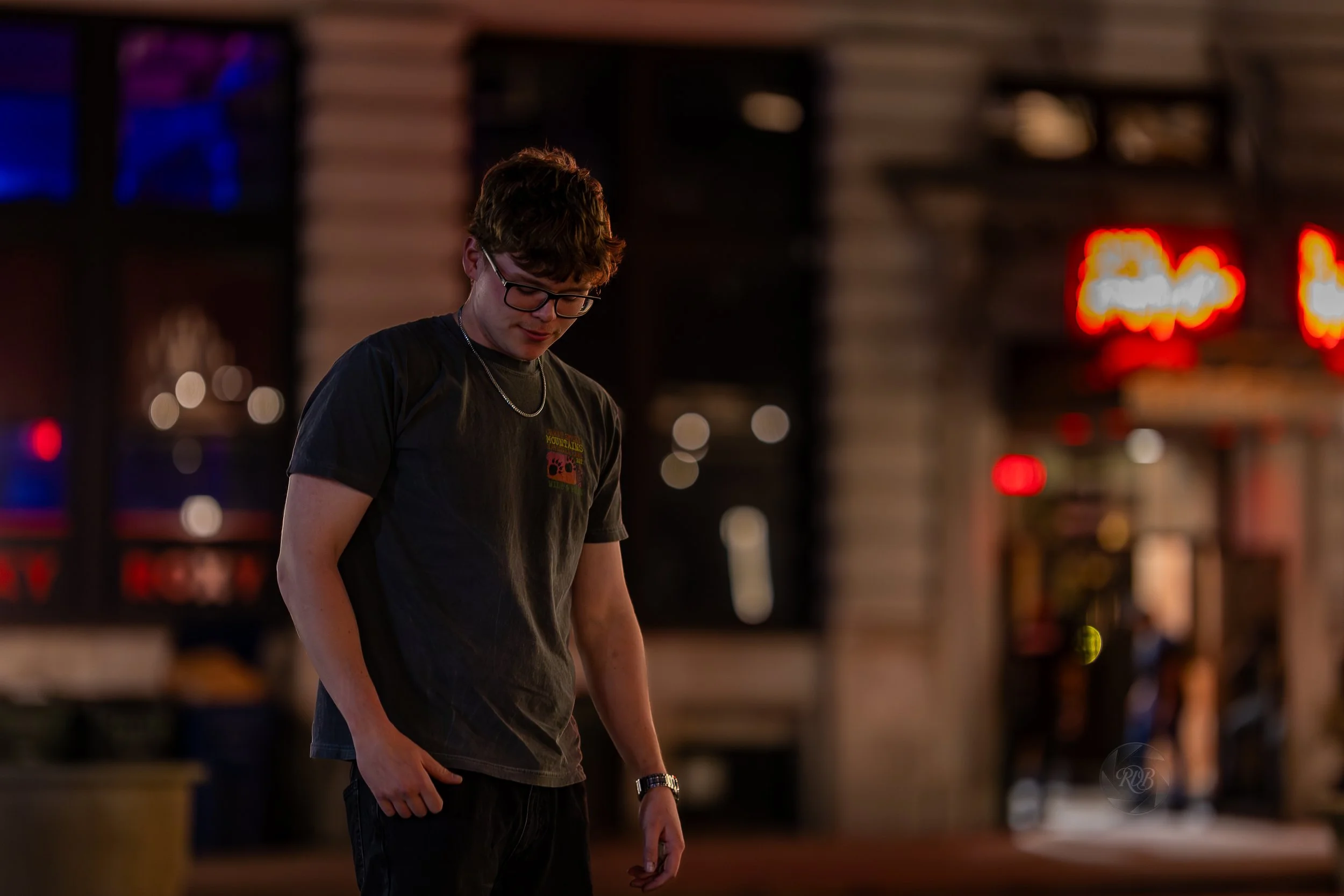 A young man with glasses, wearing a black t-shirt and a necklace, standing outdoors at night in front of a building with illuminated signs.